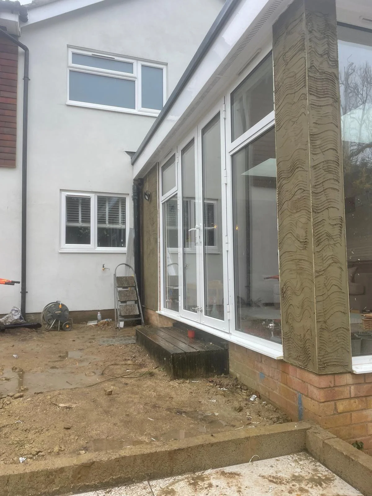 Construction site of a house with large windows and a small wooden step, construction tools and materials are visible on the ground.