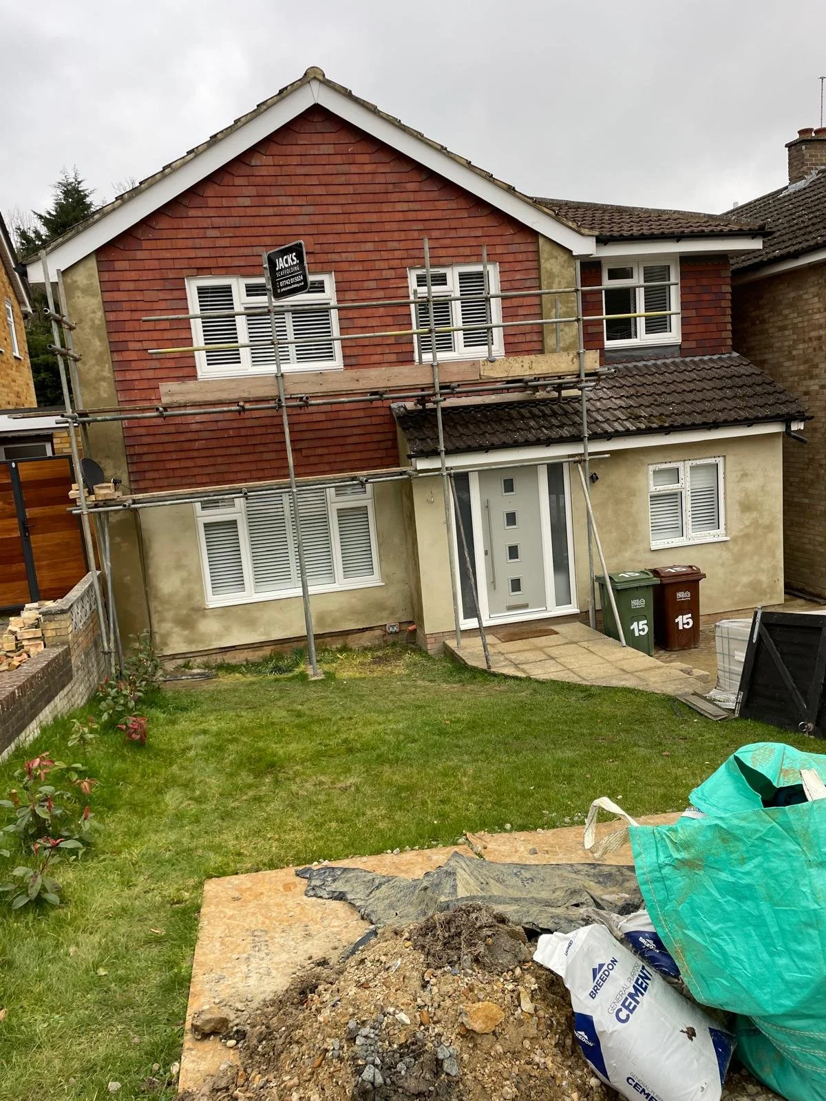 Two-story house under renovation with scaffolding, construction materials, and a small front yard with grass and a flower plant.
