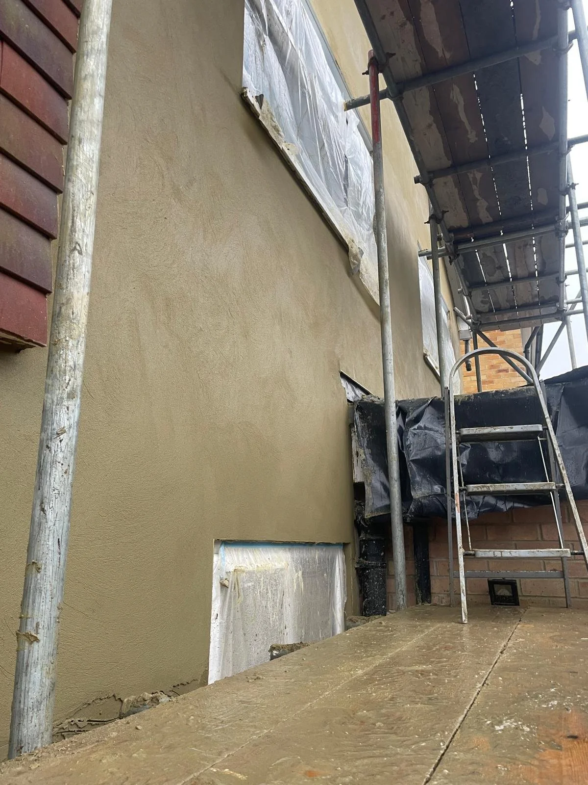 A building exterior undergoing exterior wall plastering with scaffolding. The wall is partially covered with a plastic sheet, and a small rectangular window opening is visible near the ground.