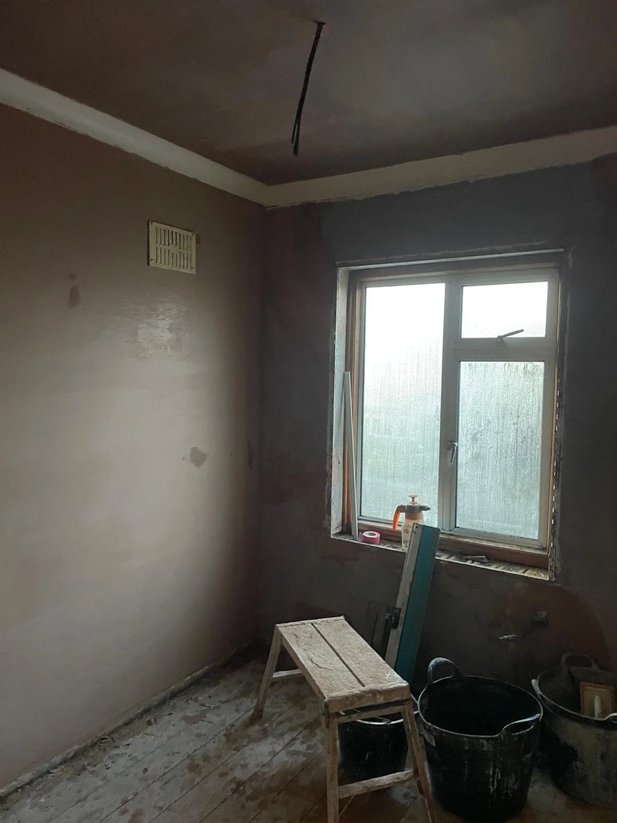 A small room under renovation with a window, construction tools, a stool, and buckets.