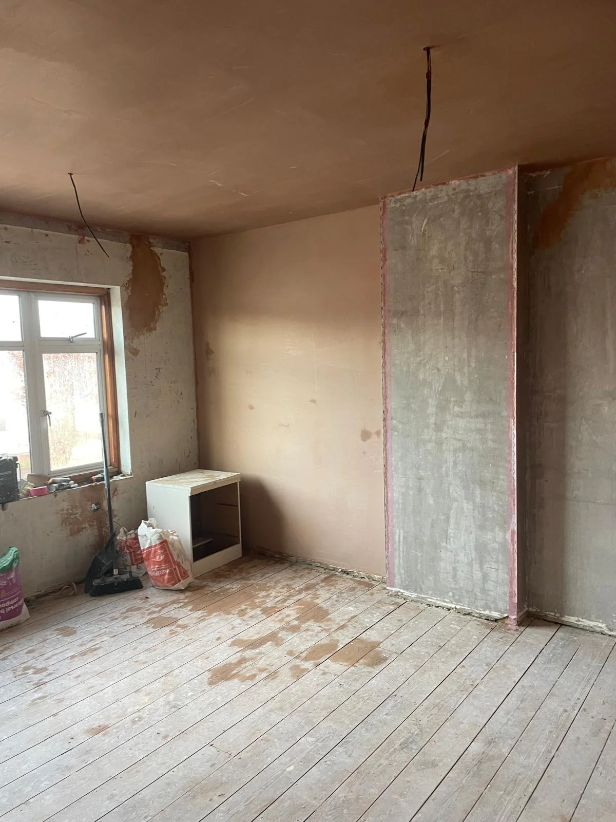 A room under renovation with unfinished walls and floor. There is a small white cabinet on the floor near a window, construction materials, and tools scattered around.