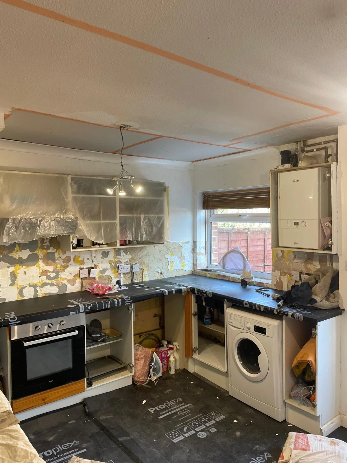 Kitchen in renovation with incomplete cabinets, a washing machine, a small oven, and insulation plastic on a wall. The ceiling has visible tape lines, and the floor is covered with protective black paper.