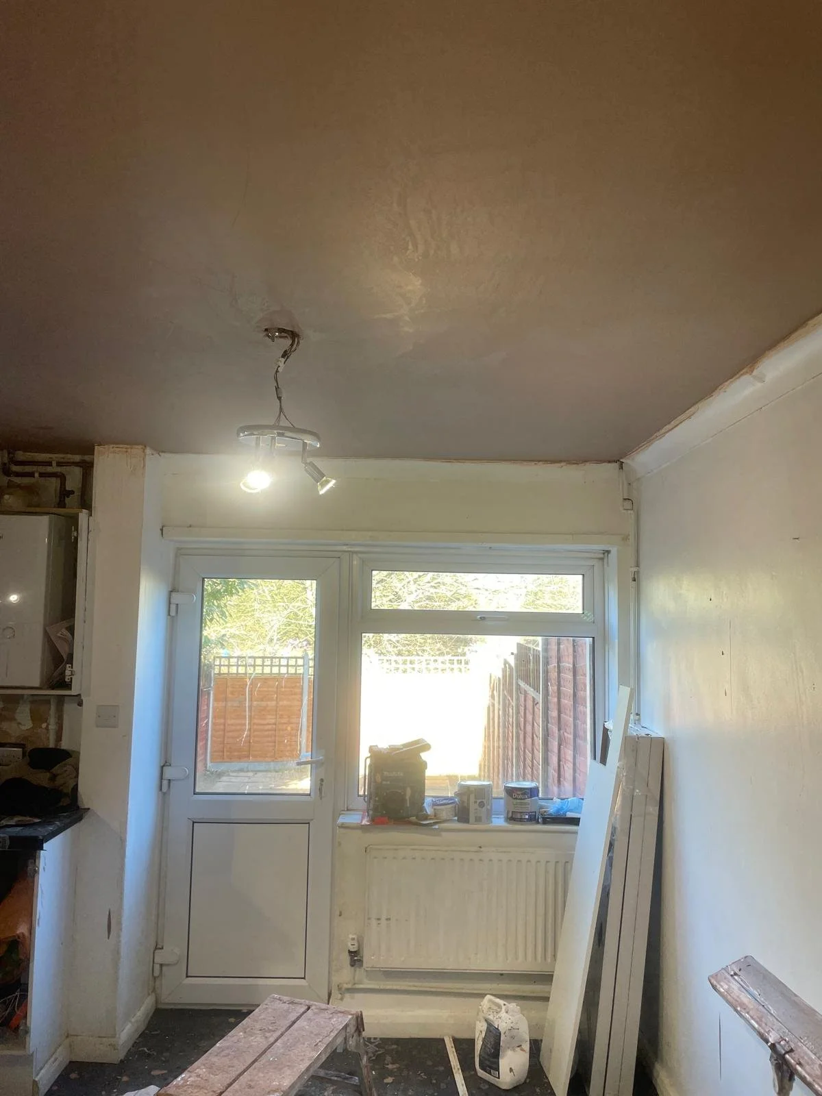 Room under renovation with a door and window looking outside, construction materials and tools on the windowsill, a stool, and a radiator below the window.