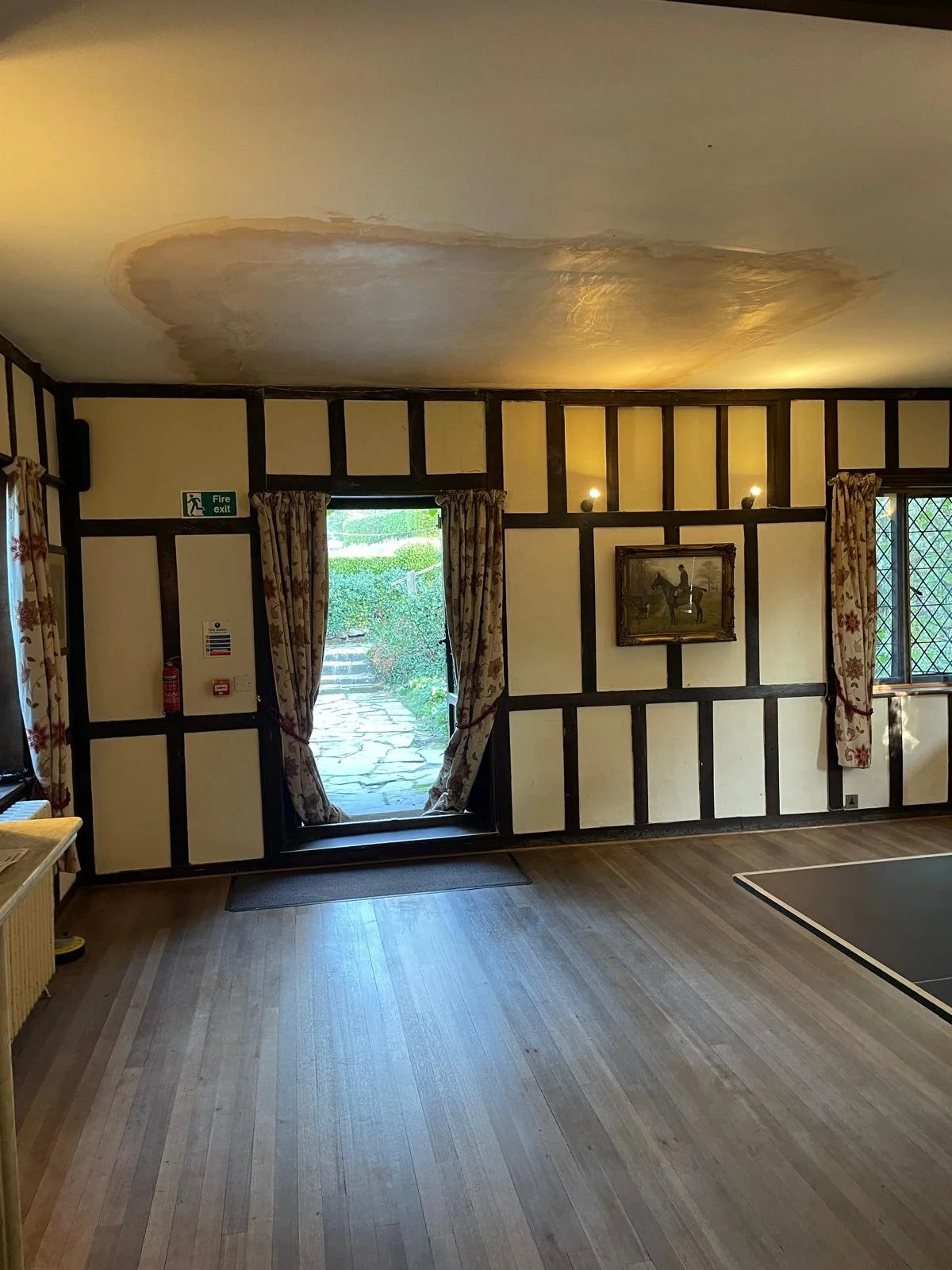 Room with beige walls, dark wooden beams, patterned curtains, a framed painting, and a damaged ceiling with a large patch of water stain.