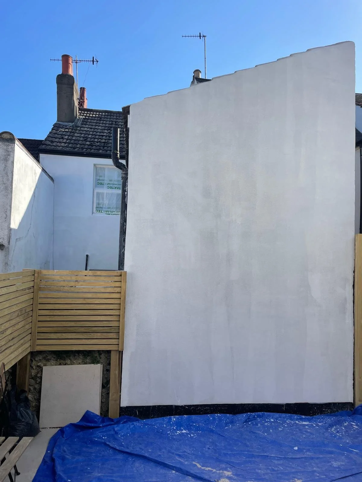 A white exterior wall of a building under renovation, with a window and a section of timber fencing, covered with a blue tarp at the bottom