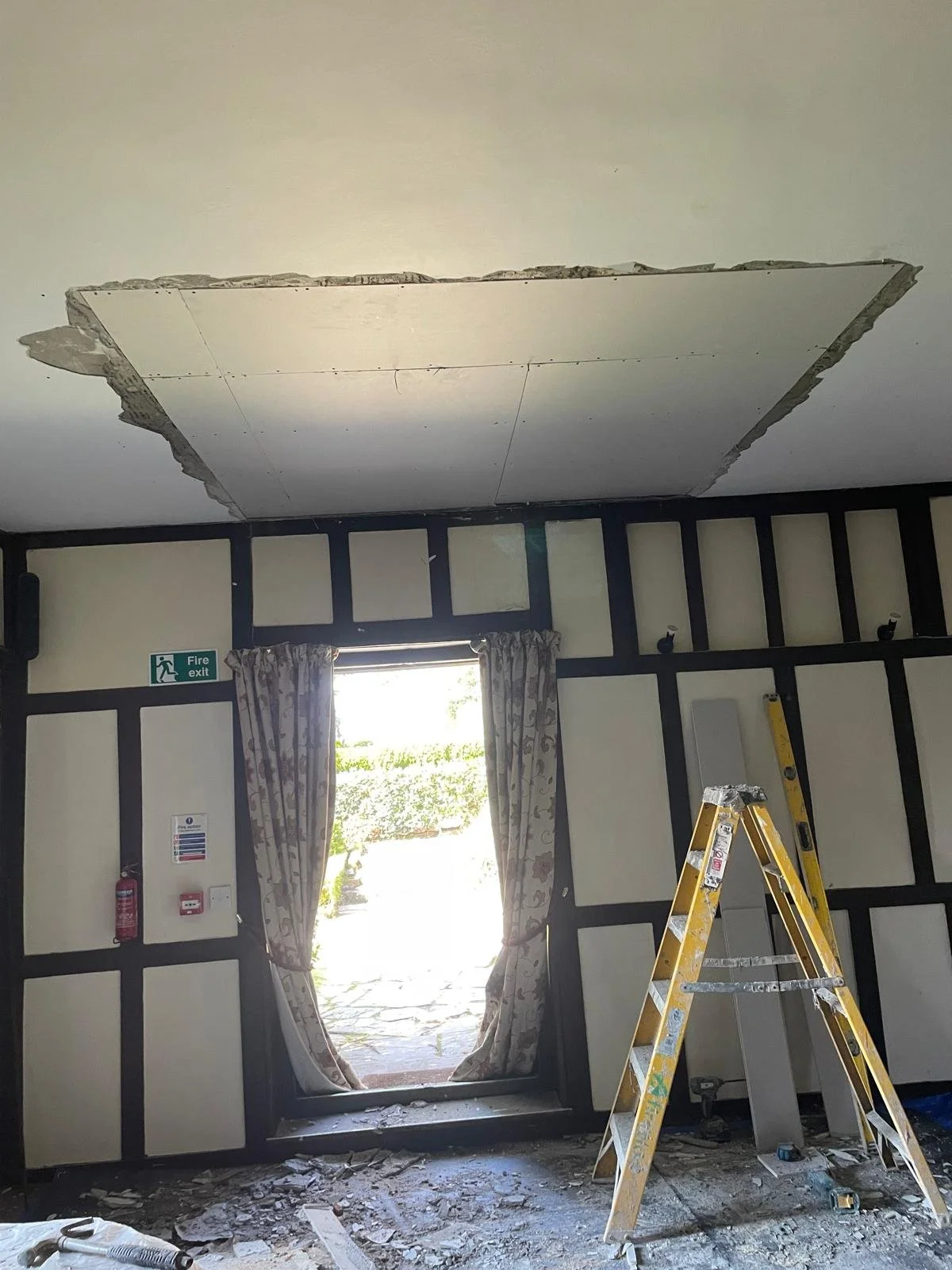 Ceiling ceiling partially removed above a window with curtains in a room under renovation, with tools and a ladder on the floor.