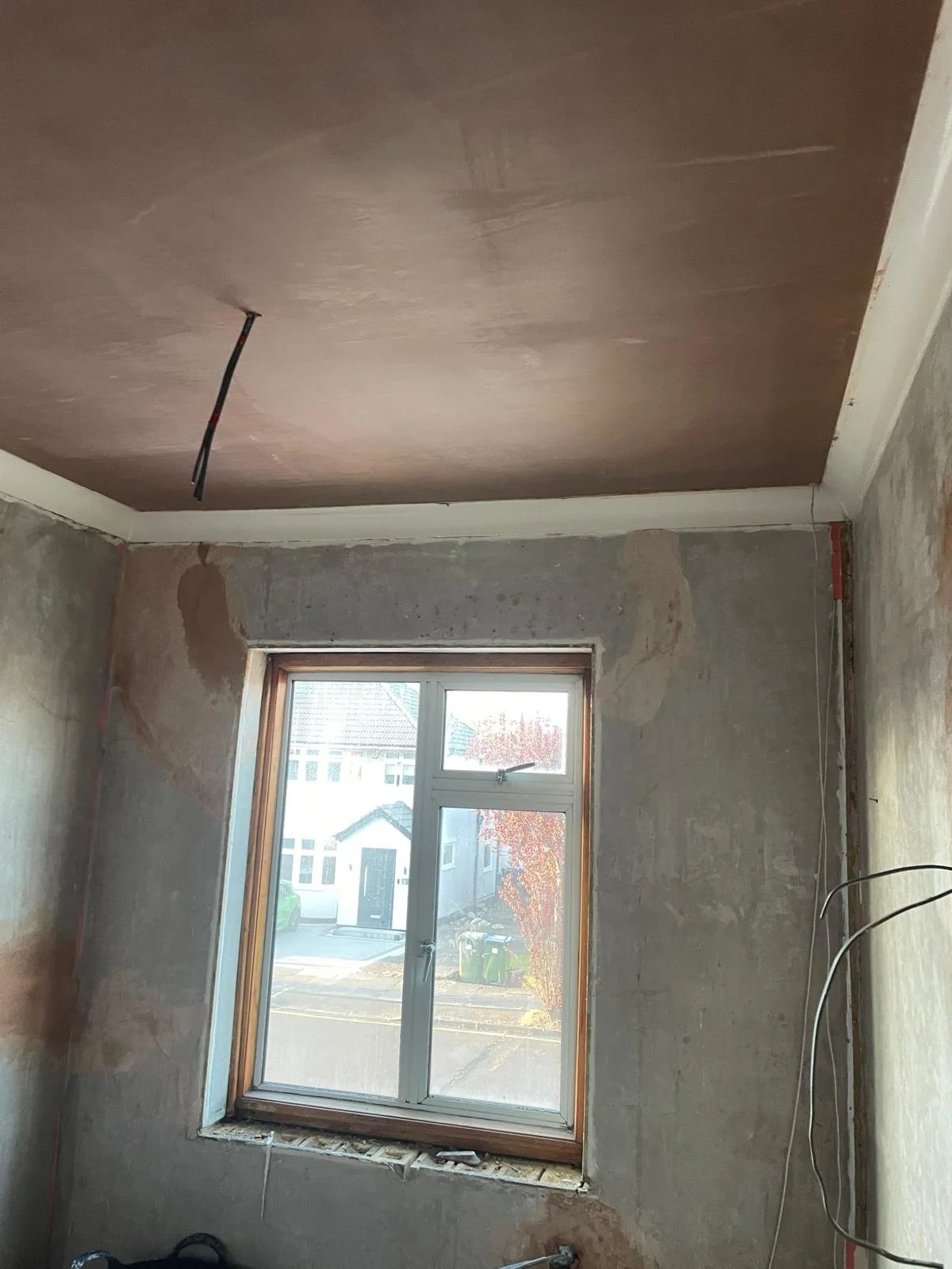 A room under renovation with an unfinished wall, a window, and a ceiling with a visible hole and wiring.