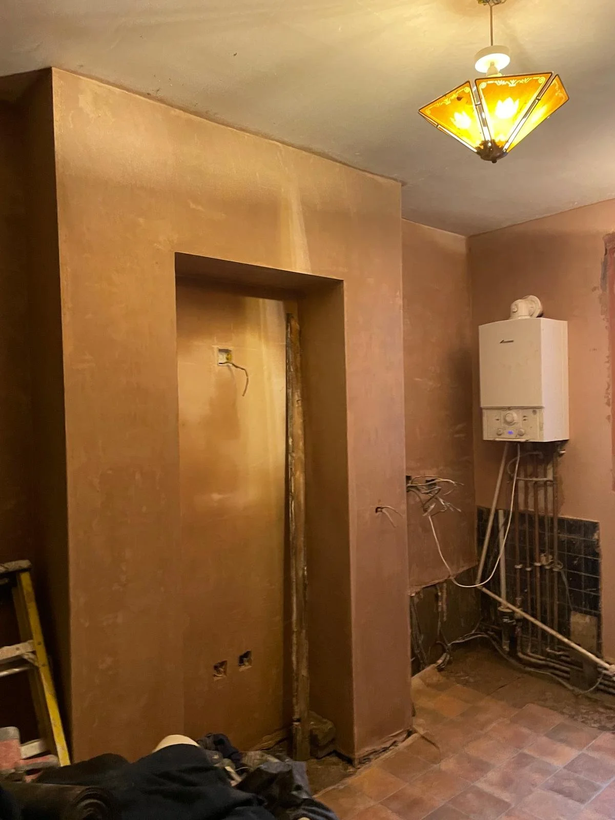 A room with unfinished walls and exposed electrical wiring, a ladder, and a boiler mounted on the wall.
