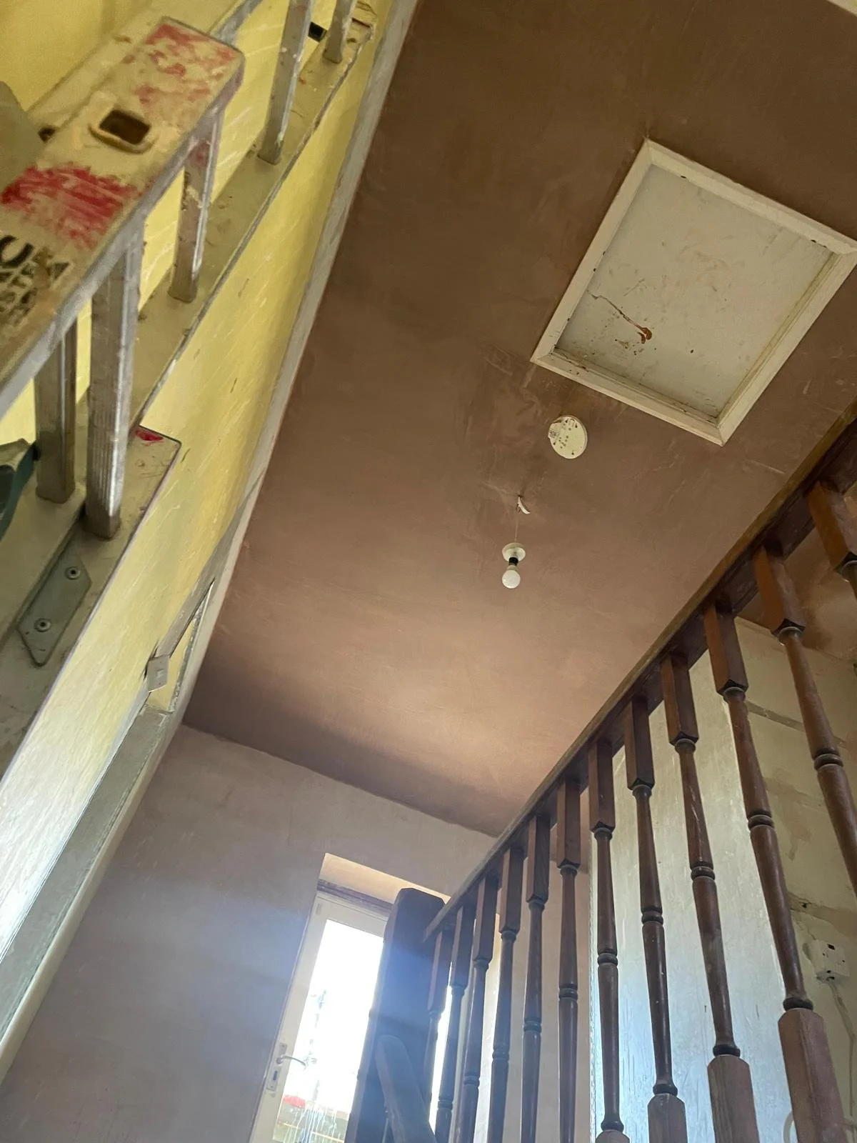 Interior view of an unfinished or renovated house ceiling with a square attic hatch, a smoke detector, and a hanging light bulb, taken from a lower angle near a staircase with wooden balusters.