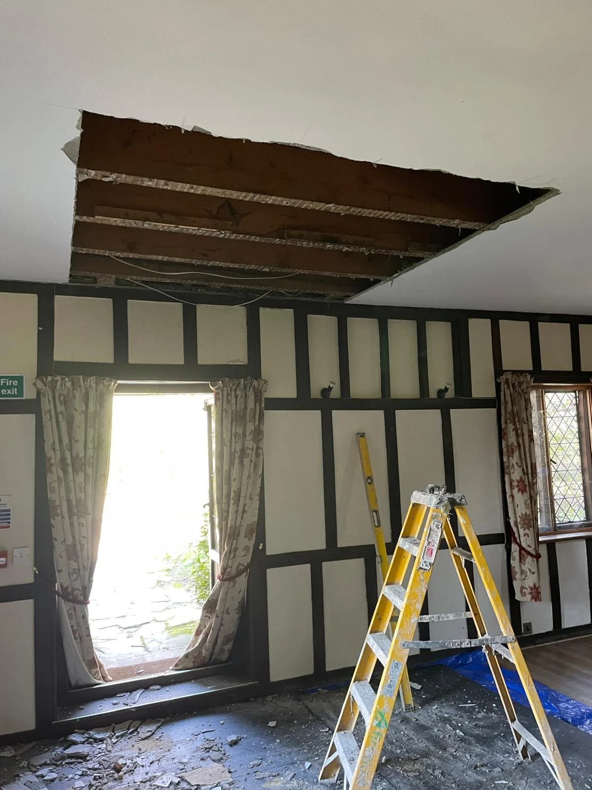 A room undergoing renovation with a large hole in the ceiling revealing the wooden beams above, two windows with floral curtains, and a yellow step ladder on a debris-covered floor.