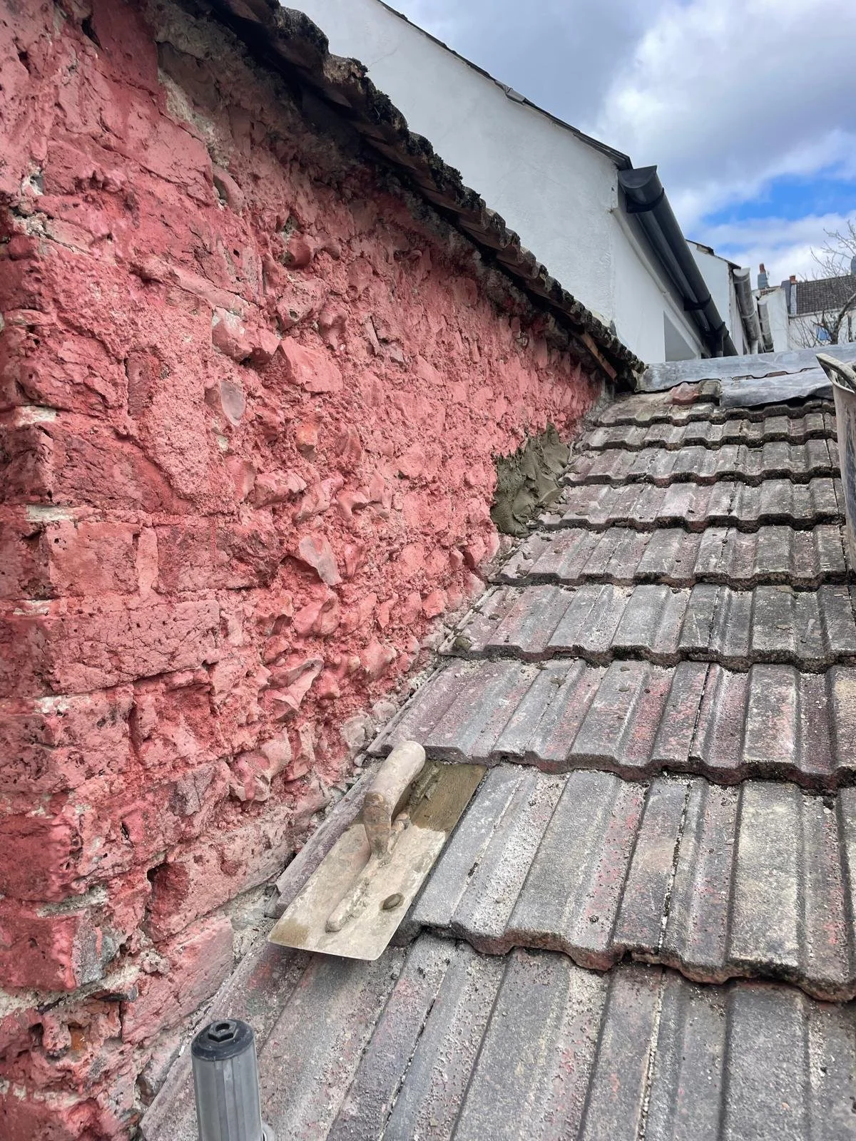 Roof repair scene showing old tiles, mortar, and a trowel near a red textured brick wall.