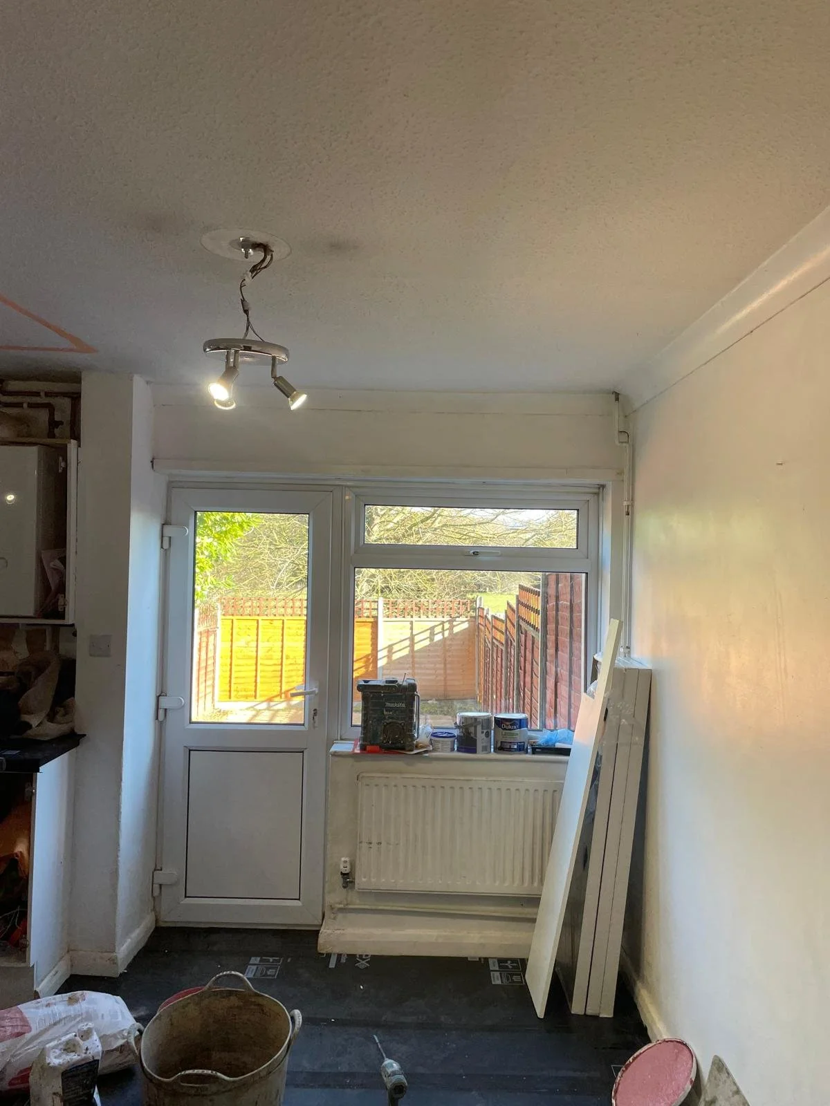 A room undergoing renovation with a window and patio door, construction materials, tools, a bucket, and a partially painted wall.