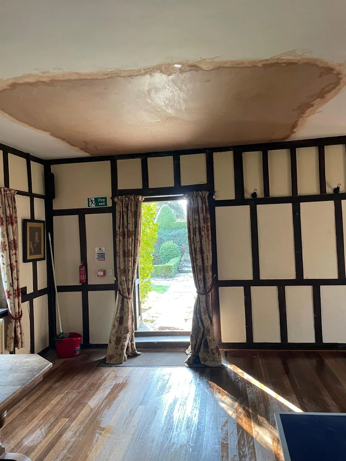 Ceiling with water damage and stain in a room with an open door leading outside, curtains, and wooden flooring.