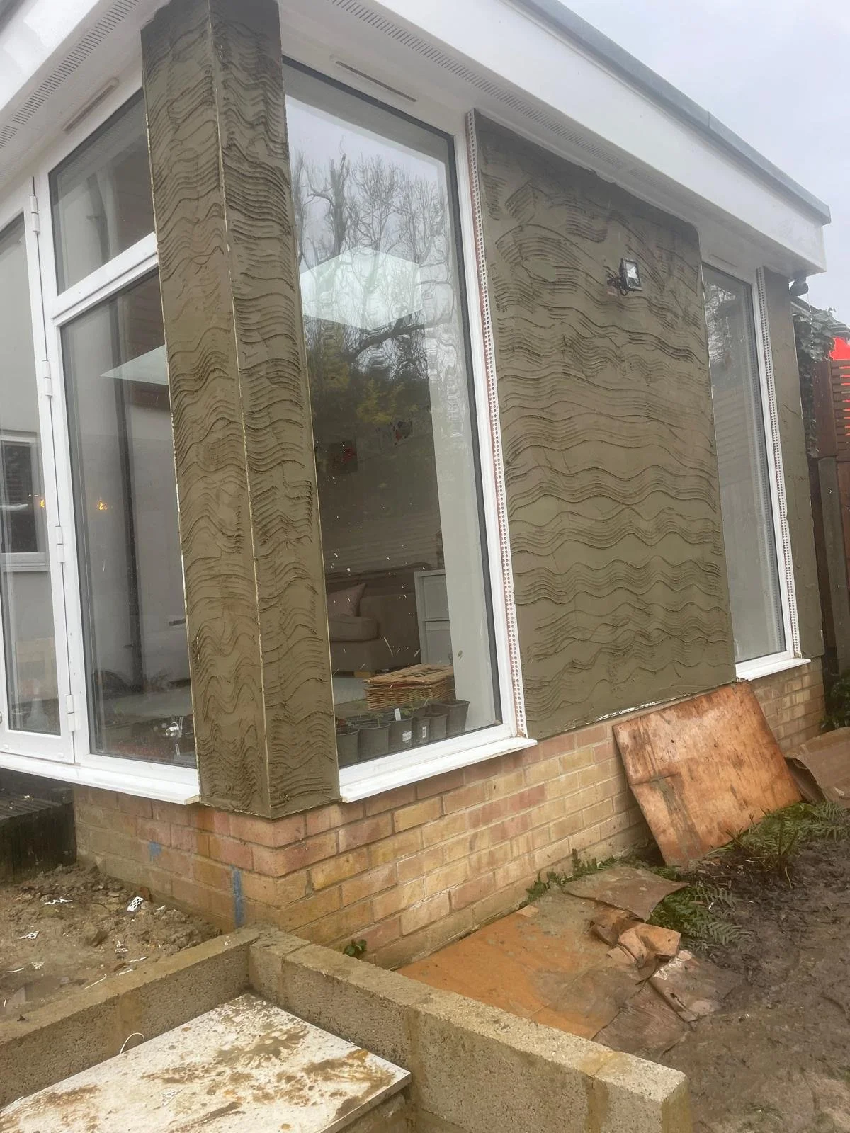 Exterior of a house under construction with large windows and textured brown siding, construction materials on the ground, and a brick foundation.