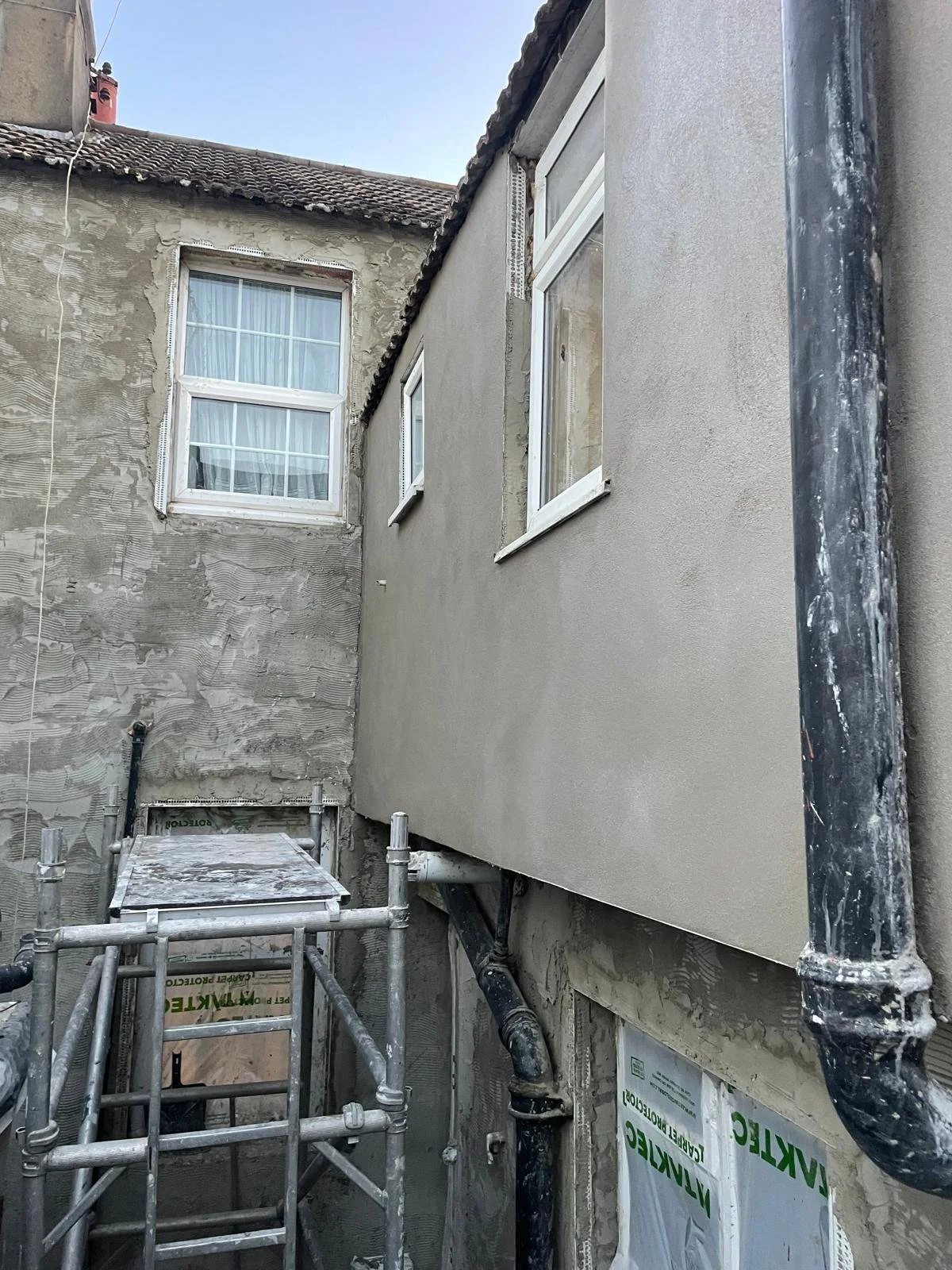 The exterior of a building under renovation with scaffolding, showing new stucco or plaster being applied to one wall and windows installed.
