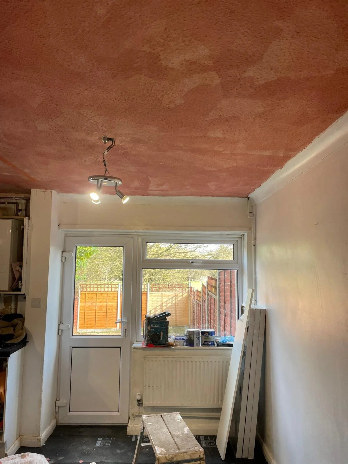 Room with window, door, radiator, and construction tools, ceiling partially painted in red, possibly during renovation.