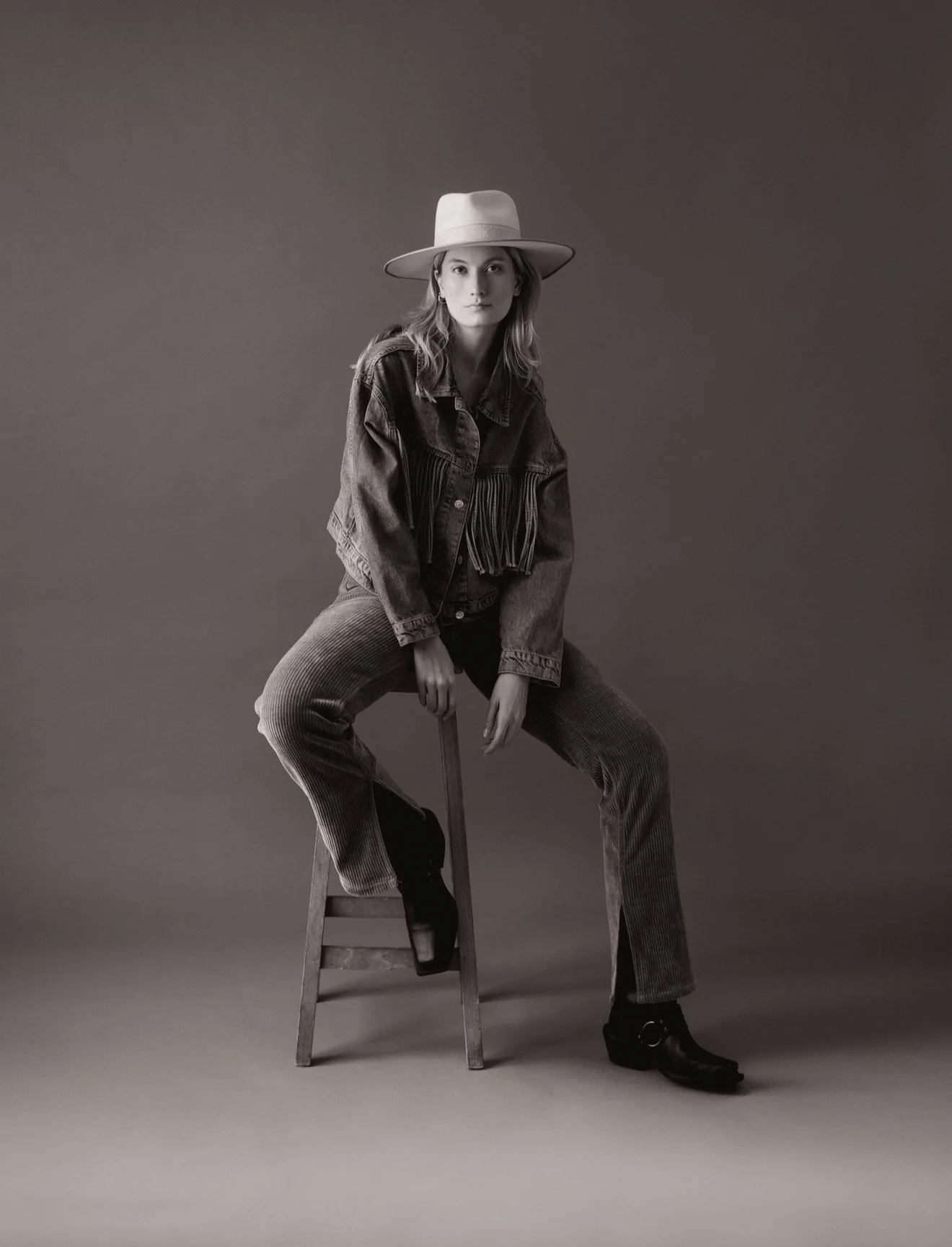 Une femme pose assise sur une chaise, portant un chapeau, une veste en jean et un pantalon en velours côtelé, en noir et blanc.