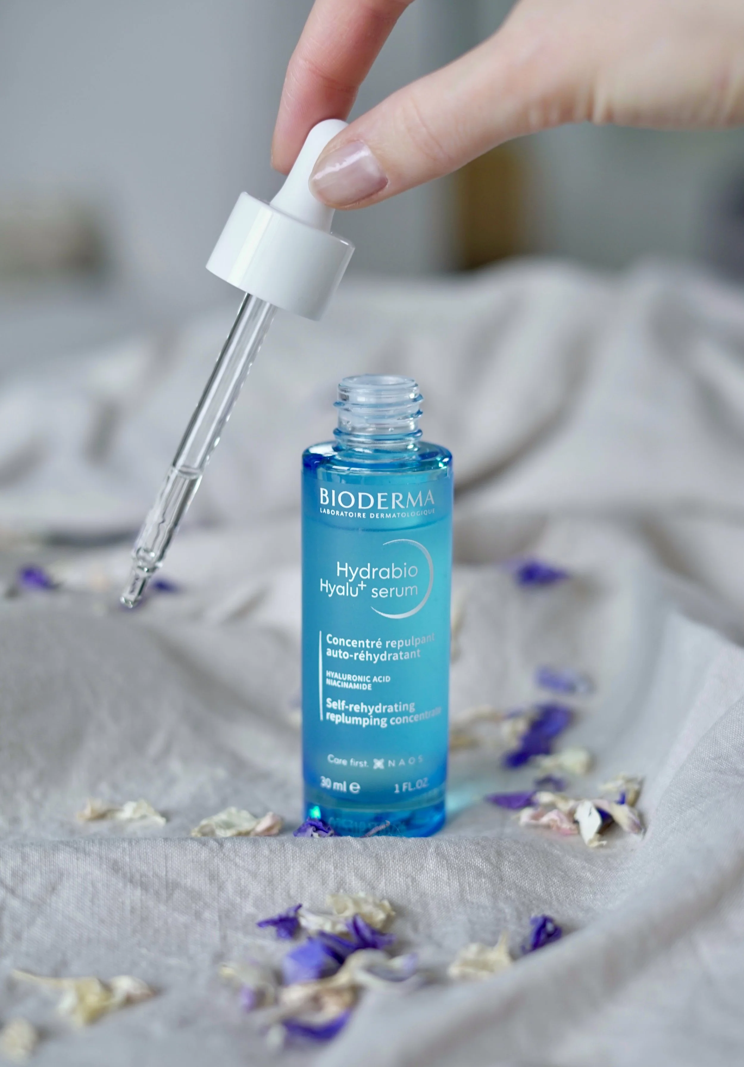 Une bouteille de sérum hydratant Bioderma Hyalu+ sur un tissu blanc avec des fleurs séchées autour, une main tenant une pipette prête à appliquer le produit.