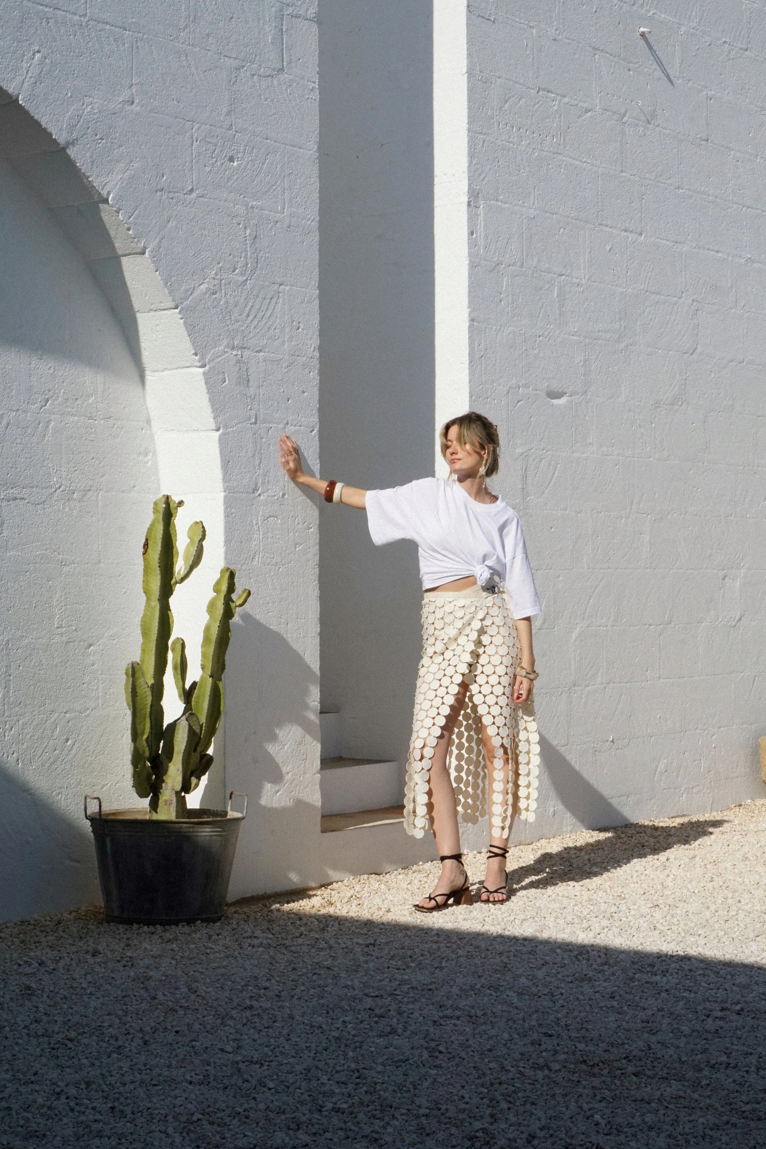 Une femme en robe beige avec des cercles blancs, portant des talons noirs, se tient dans une cour ensoleillée contre un mur blanc avec un arbre cactus en pot à côté d'elle.