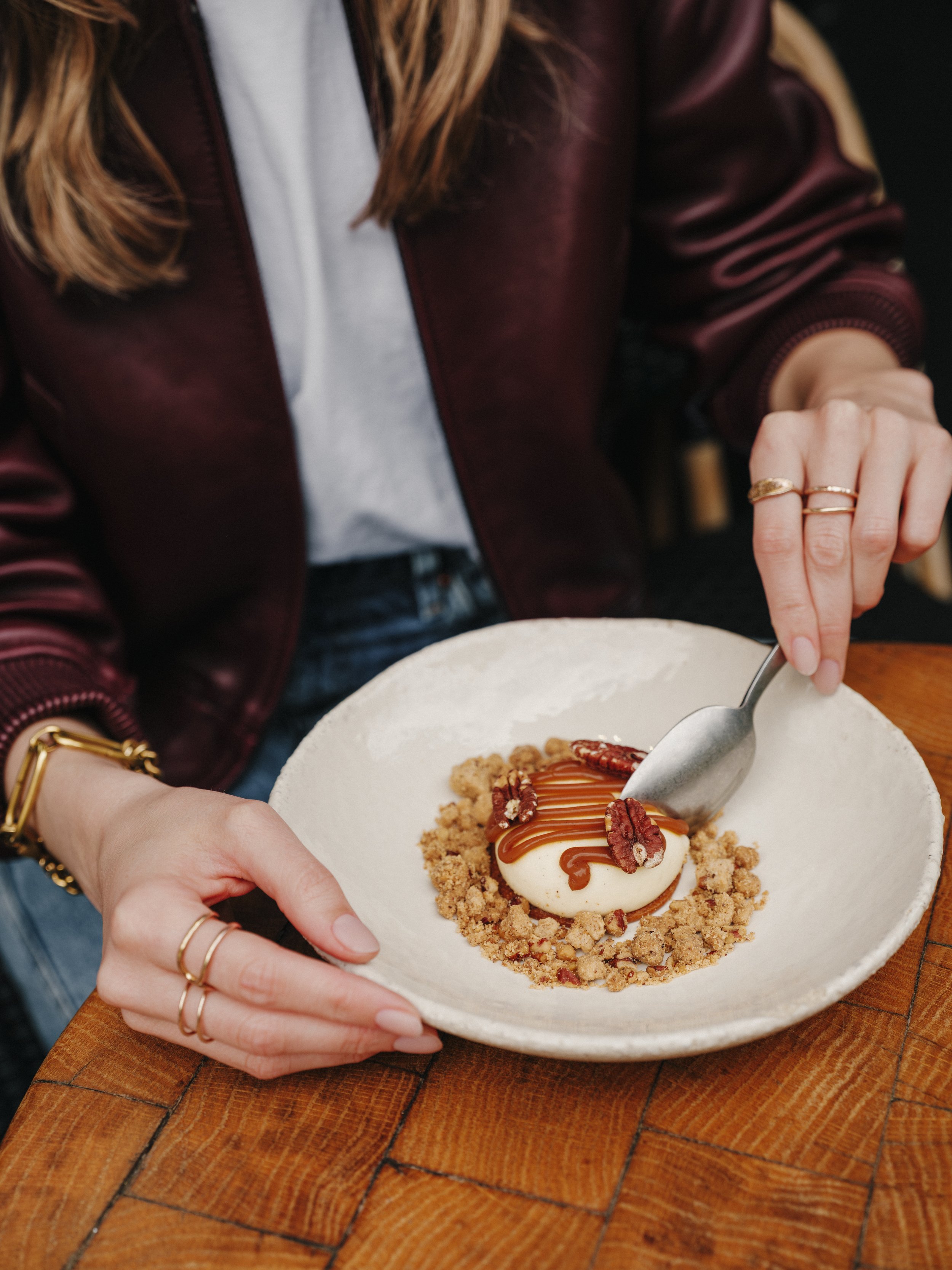 Une personne tient une assiette avec un dessert à la crème, décoré de caramel, de noix de pécan et de crumble, sur une table en bois.