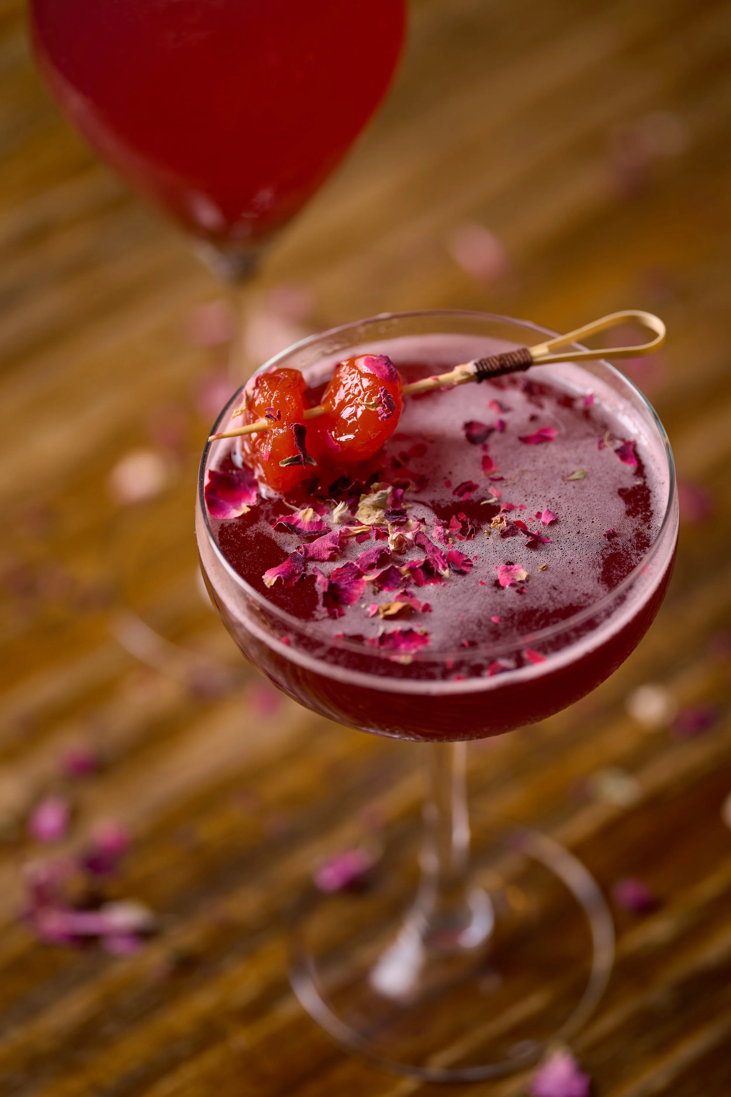 Un verre de cocktail rouge avec des pétales de rose et une cerise comme garniture, placé sur une surface en bois avec des pétales de rose dispersés autour.