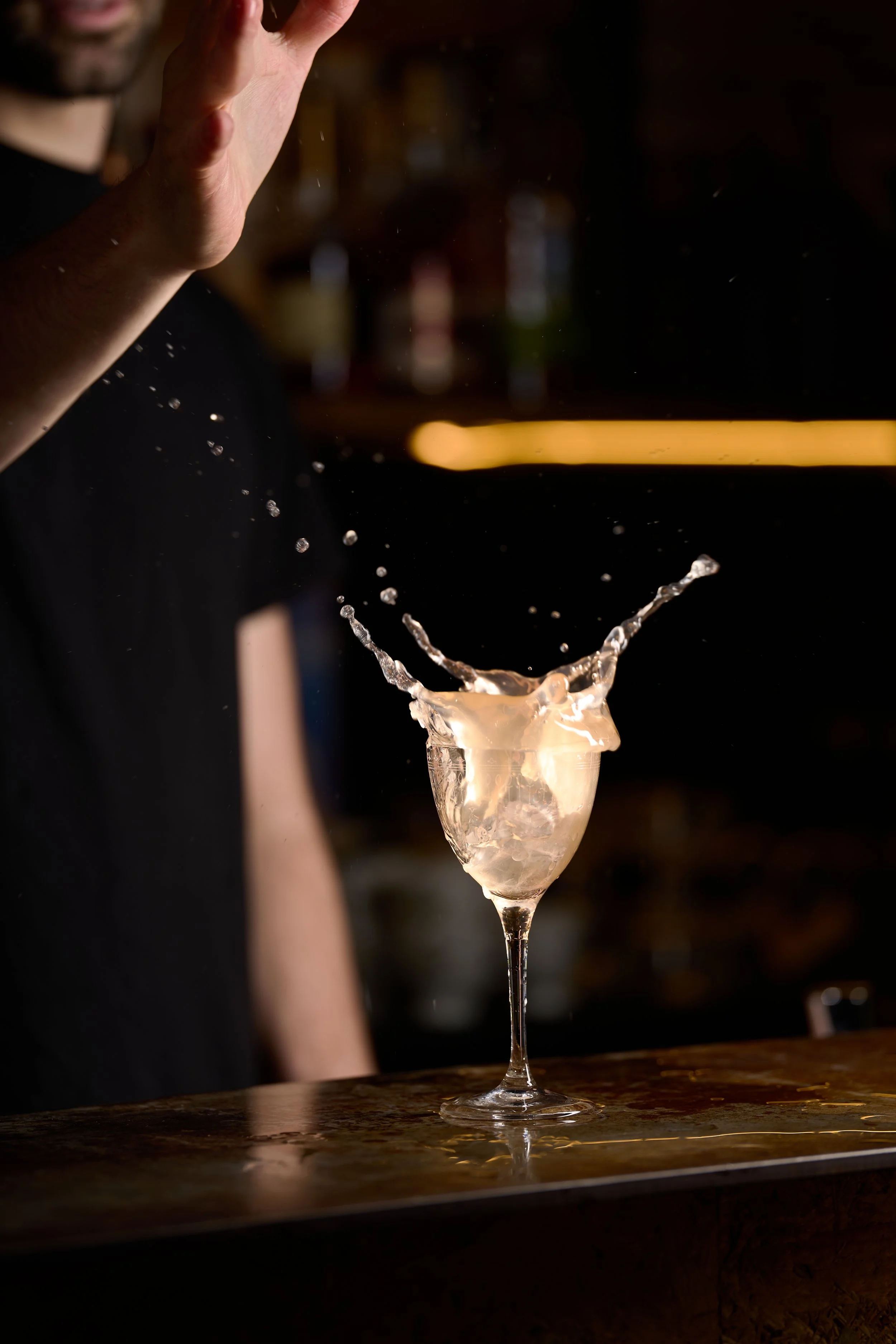 Un cocktail dans un verre à vin, avec une explosion de liquide à la surface, dans un bar sombre.
