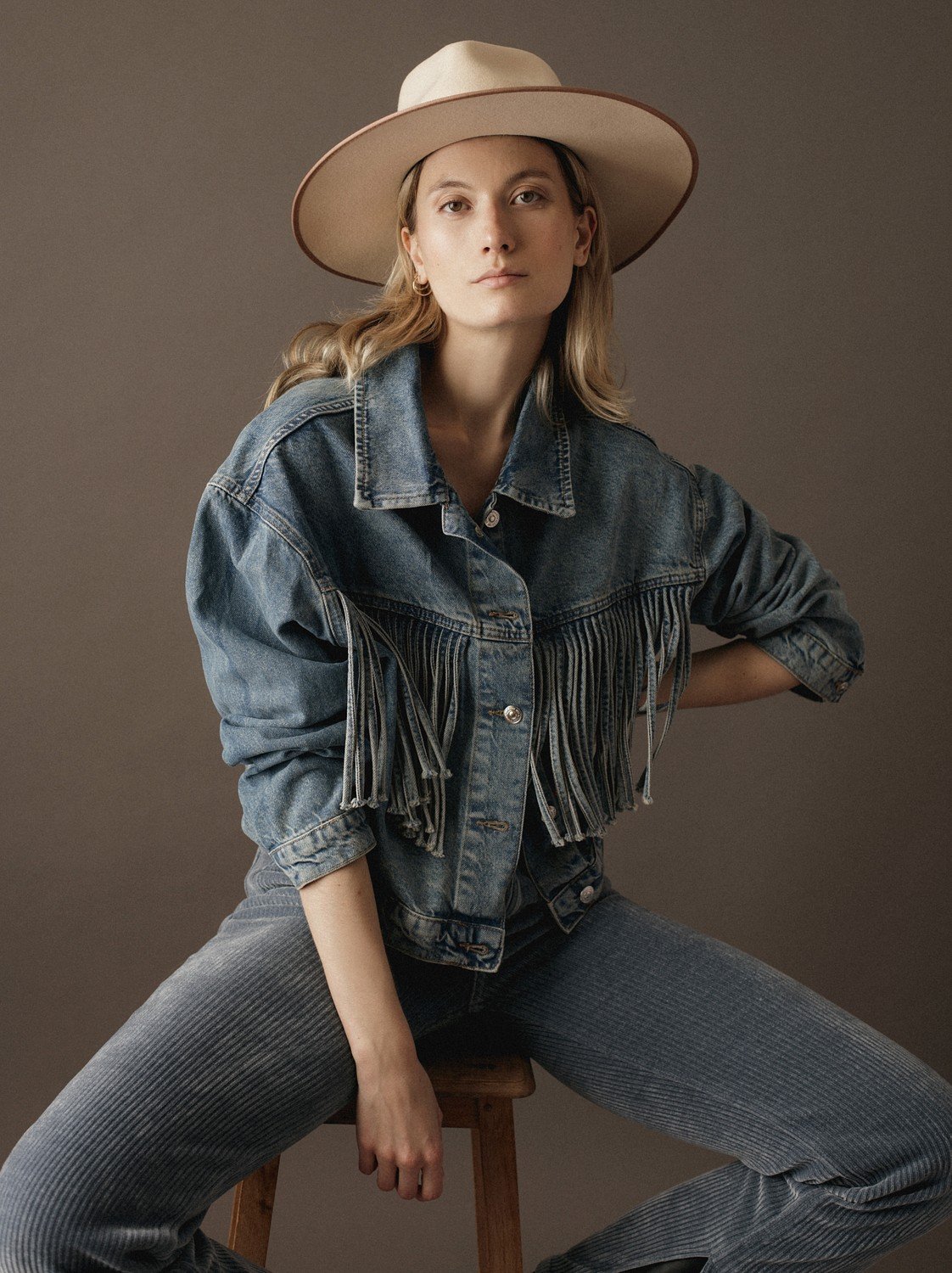 Femme portant un chapeau beige, veste en denim à franges, pantalon en velours côtelé gris, pose assise sur une chaise en bois, fond neutre.