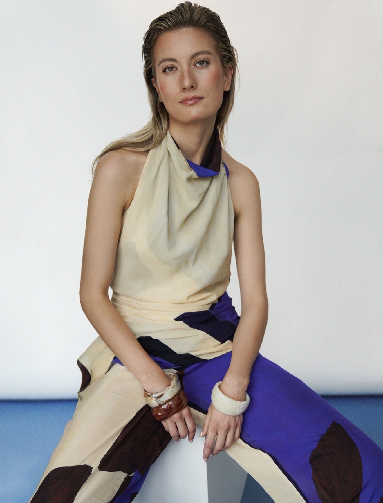 Une femme blonde à l'air confiante, portant une robe sans manches beige avec des accents bleus et violets, assise contre un mur blanc, les mains posées sur ses cuisses, portant plusieurs bracelets en bois et en pierre.