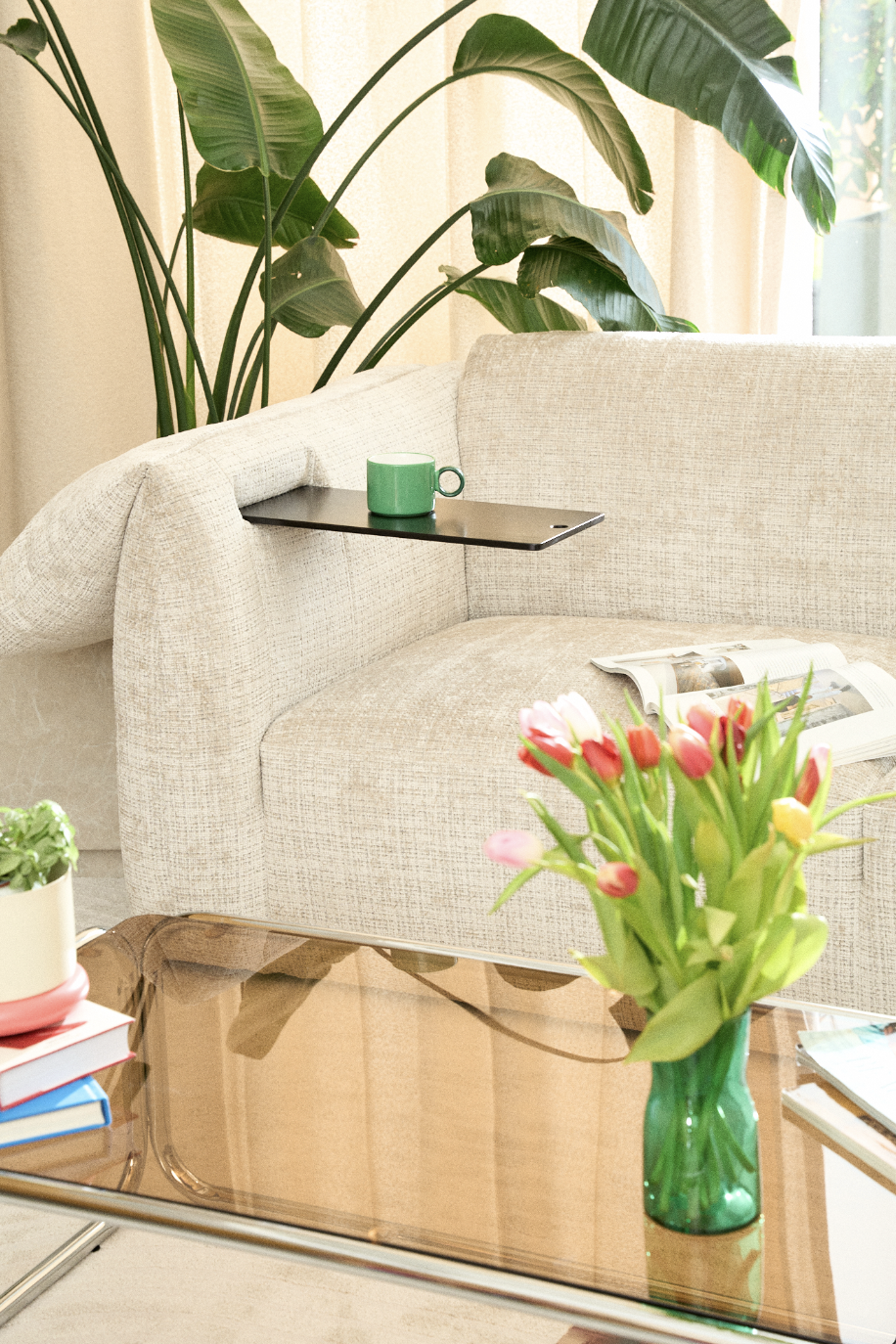 Un salon avec un canapé beige, une plante verte en arrière-plan, une table en verre avec un vase de tulipes roses et rouges, une tasse verte sur une tablette noire appuyée sur le canapé, une lampe de lecture à côté avec un magazine ouvert.