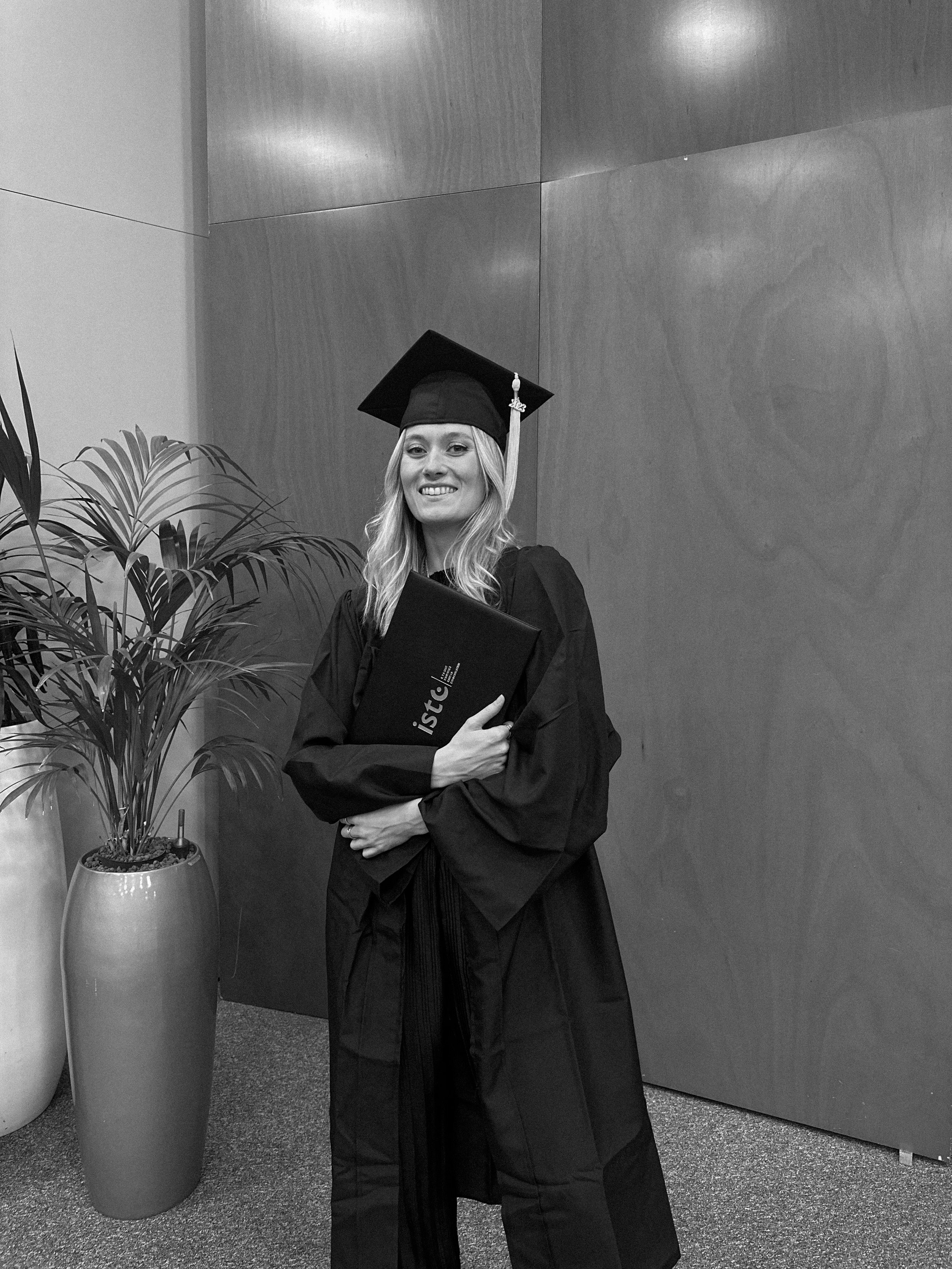 Une femme habillée en robe de cérémonie et portant une toque de diplômé, tenant un diplôme, souriant, en intérieur avec un mur en bois et un grand pot de plante à côté.