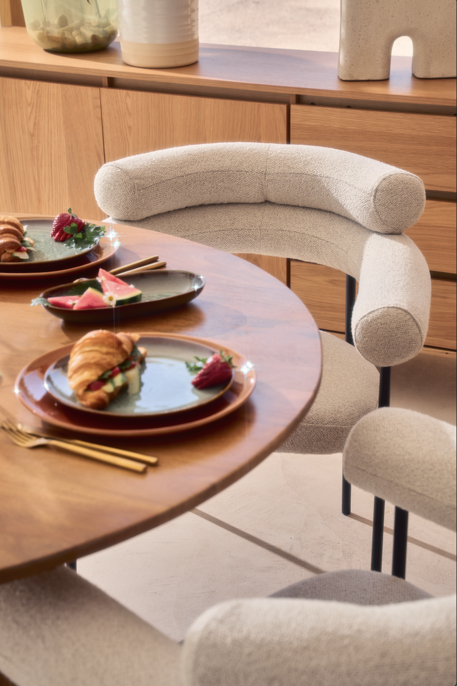 Vue d'une table avec des assiettes de nourriture, notamment des croissants, des fruits et des pâtisseries, accompagnée de chaises modernes dans une salle à manger chaleureuse.