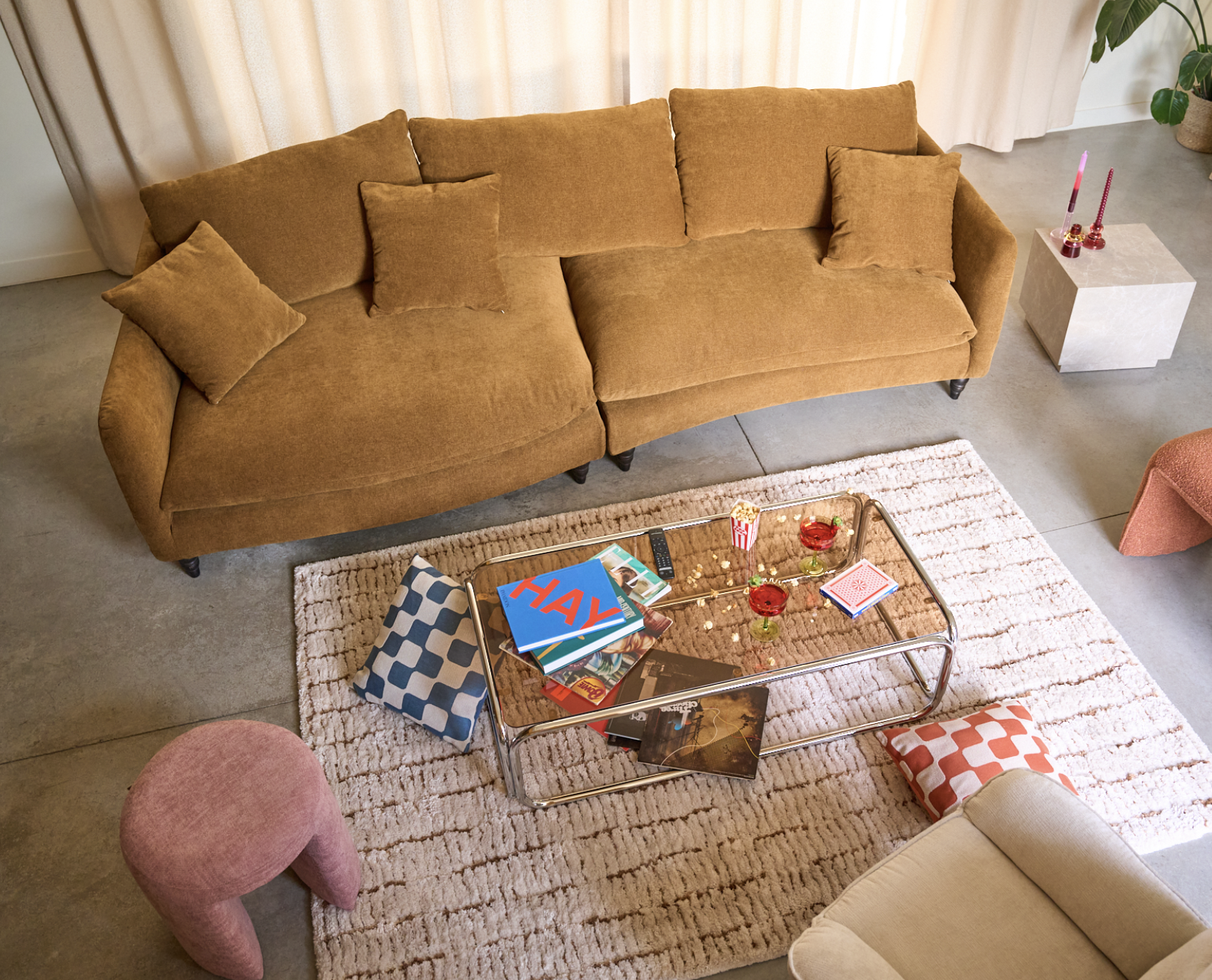 Un salon avec un canapé marron, une table basse en verre avec des magazines et des boissons rouges, un coussin à motifs rouges et blancs, des coussins marrons, un pouf rose, un tapis beige, une petite table avec des bougies roses, et une plante verte en arrière-plan.