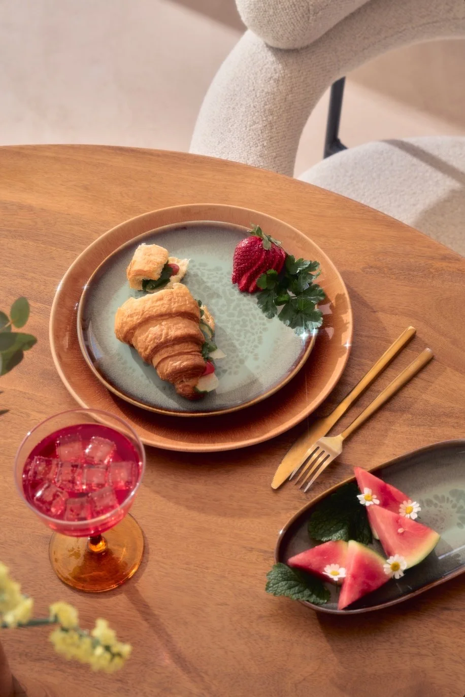 Assiette avec un croissant, une fraise et des feuilles de menthe, accompagnée de tranches de pastèque décorées de petites fleurs, et un verre de boisson rose avec des glaçons, sur une table en bois.