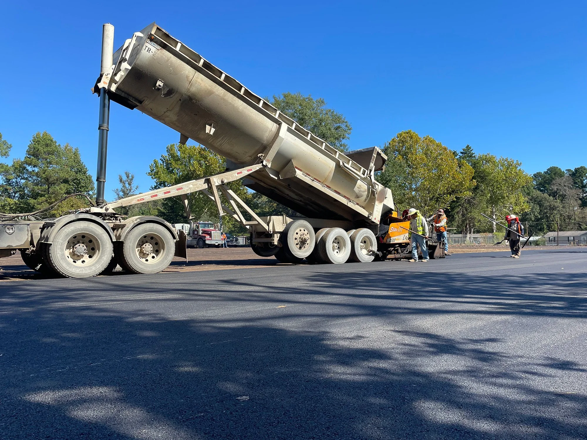 A dump truck unloads asphalt onto a road as workers in safety vests supervise the paving process under clear skies.