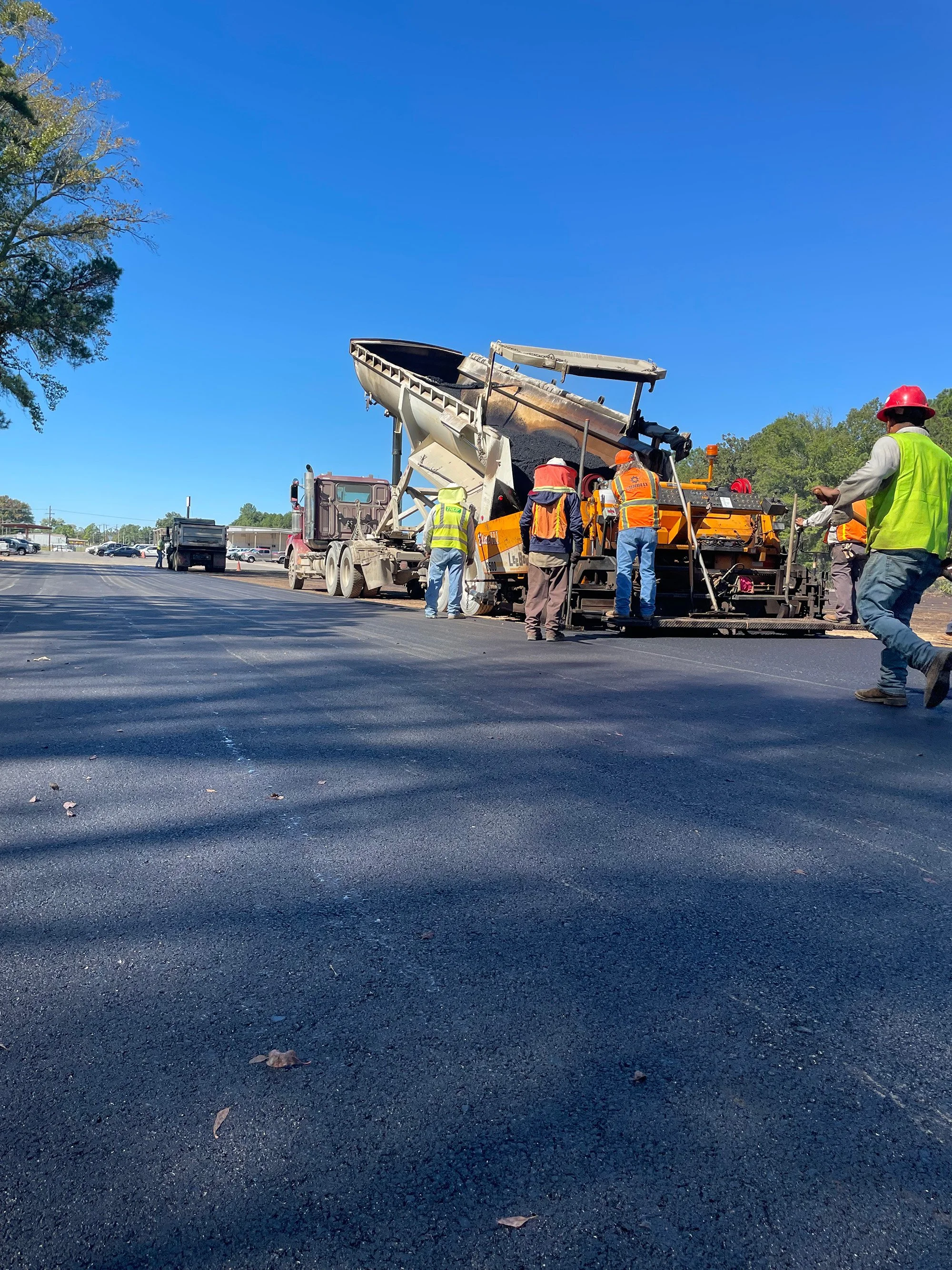 A dump truck is angled to release asphalt onto a road, with safety-vested workers monitoring the paving work in East Texas.