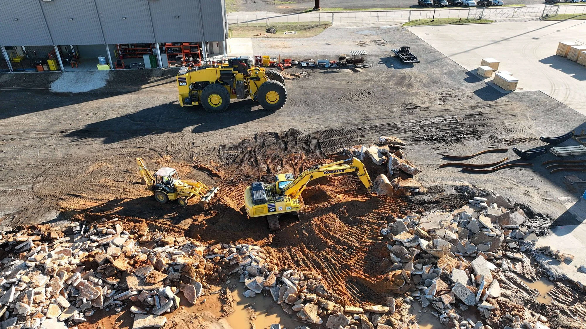 A yellow excavator moves rocks while a loader works nearby on a construction site with machinery and debris visible in the background.