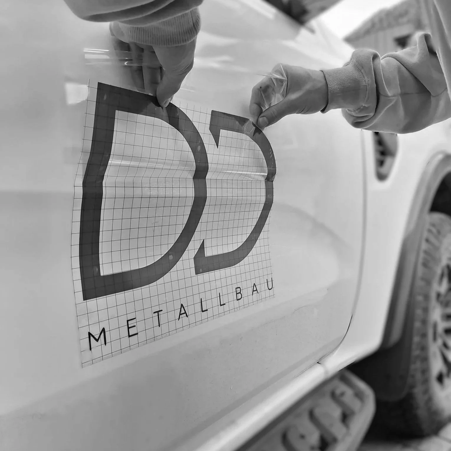 Person klebt Metall-Logo mit der Aufschrift 'METALLBAU' auf die Seite eines Fahrzeugs
