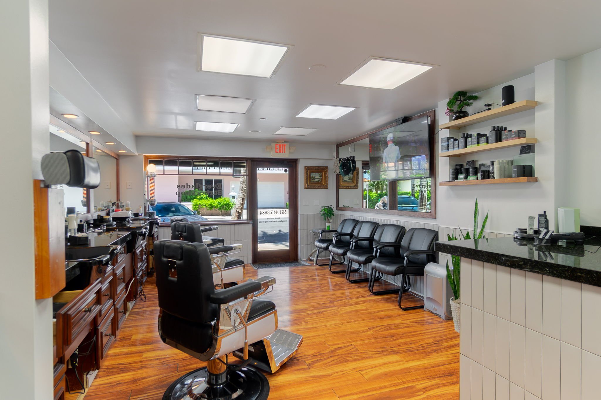 Interior of a barbershop with black chairs, wooden cabinets, a reception counter, a TV, and large windows allowing natural light.