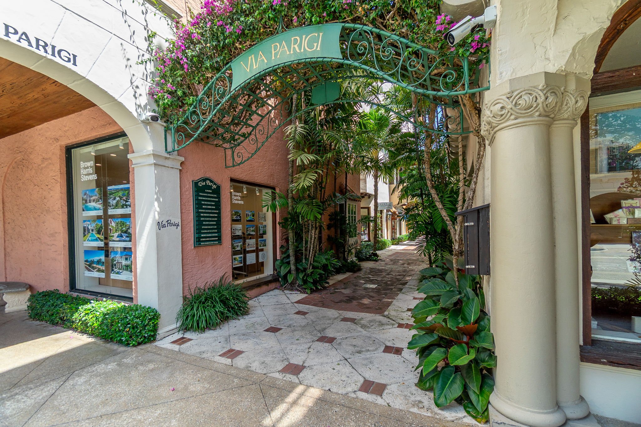 An arched entrance with green ornate metalwork and a sign reading 'VIA PARIGI' over a lush, tropical garden pathway, flanked by pink and cream-colored buildings with display windows.