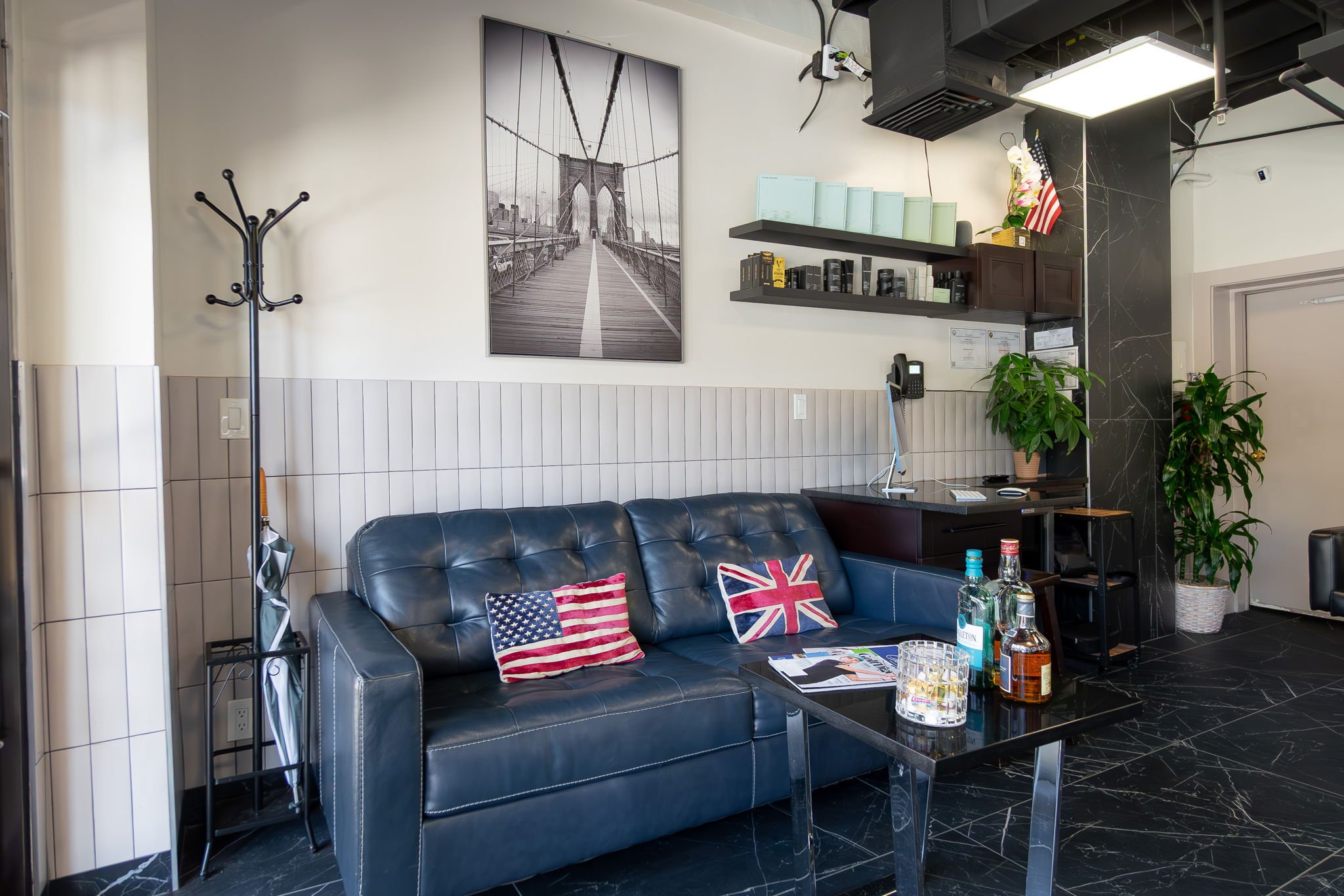 Lobby area with a black leather couch decorated with American and British flag pillows, a black coffee table with bottles and glasses, a corner desk with a computer phone, wall shelves with books and decor, a large black and white bridge photo on the