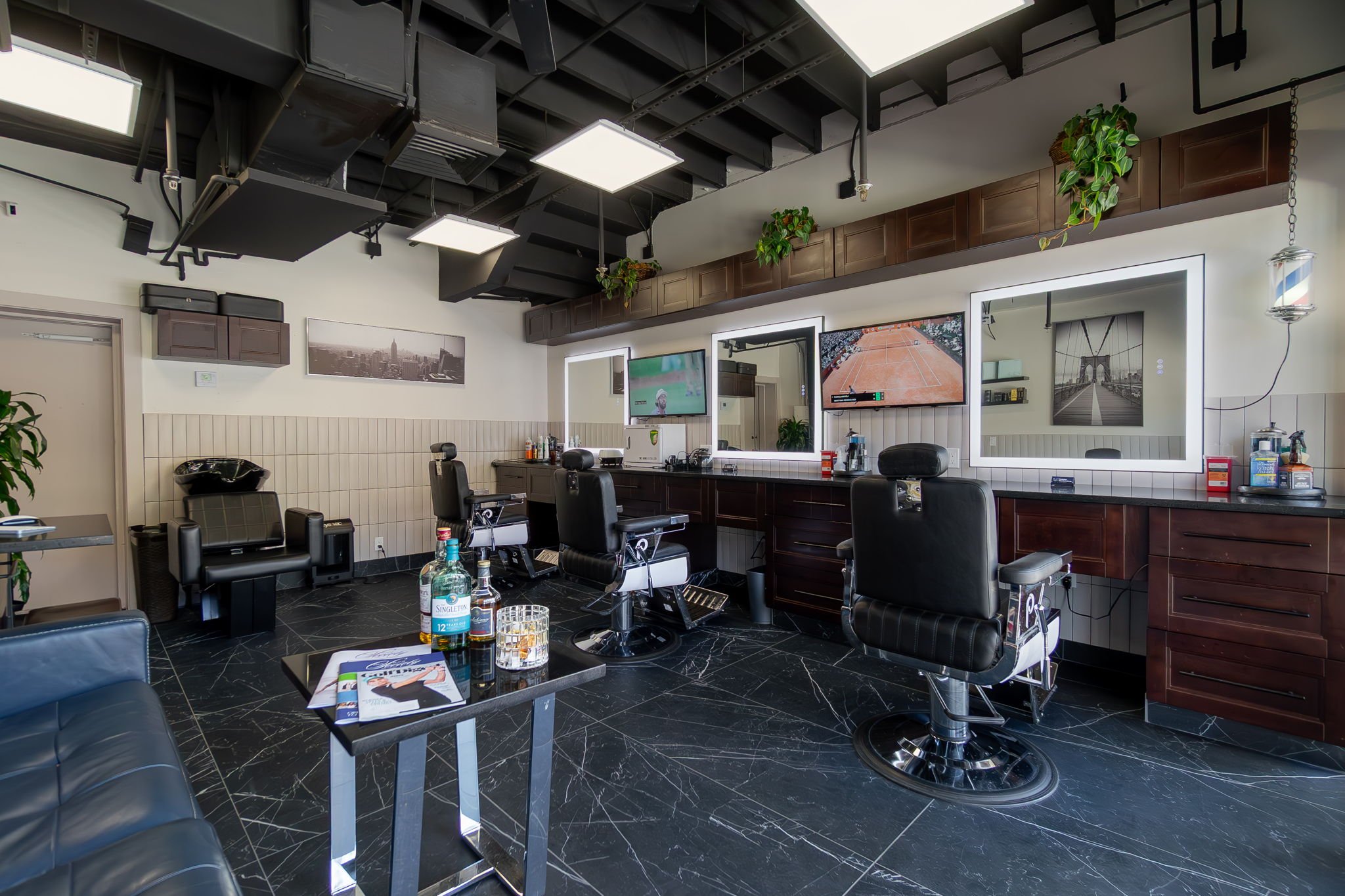 Barber shop with three barber chairs, large mirrors with built-in lighting, and a television displaying a tennis court. The shop has black marble flooring, wall art, plants, and a black leather sofa in the foreground.