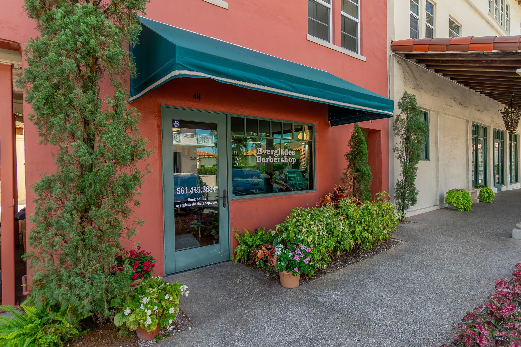 Exterior of Everglades Barbershop with green door, large window, blue awning, potted plants, and shrubs in front.