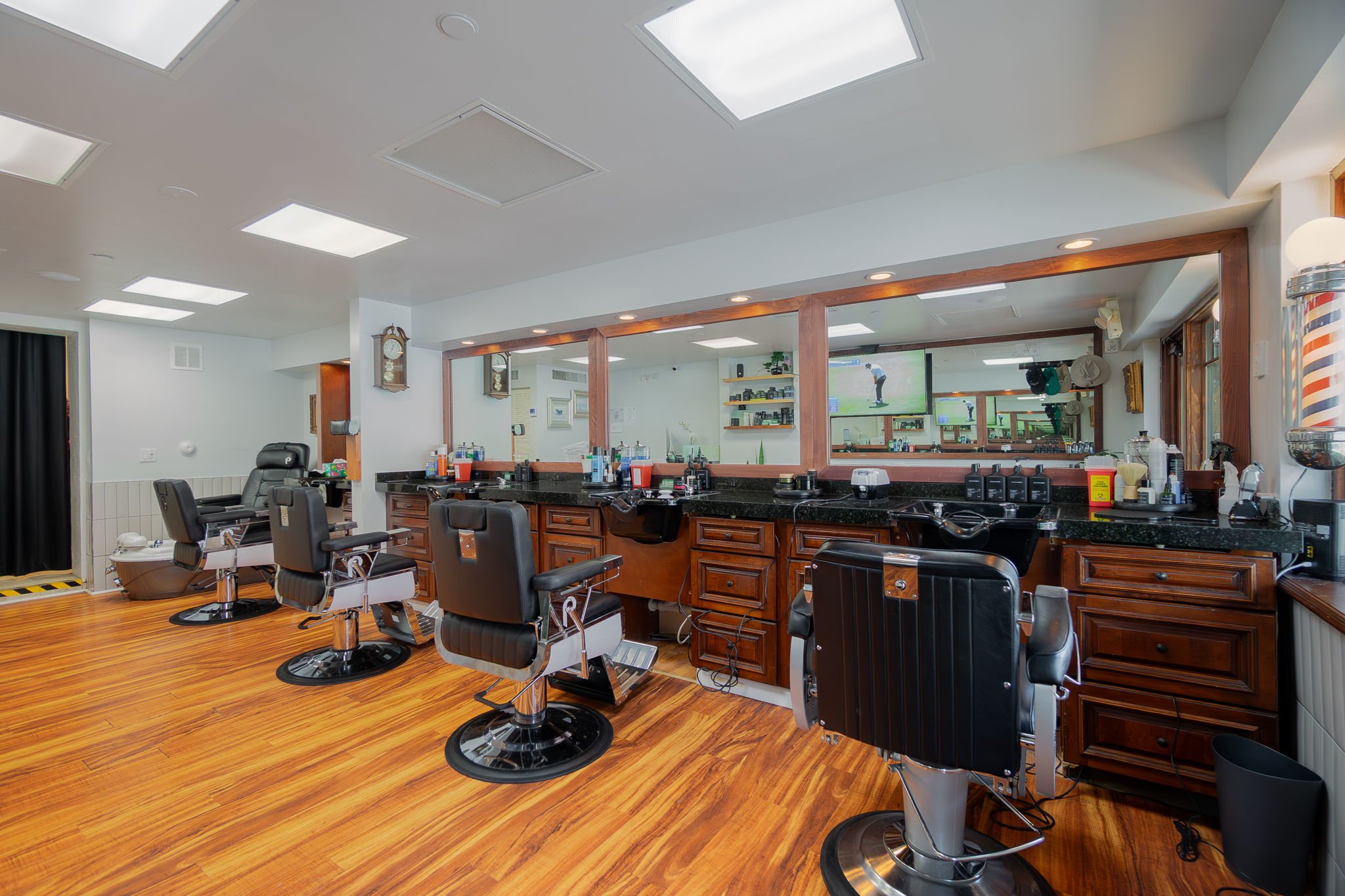 Barber shop with four black chairs in front of a wooden counter with mirrors, hair cutting tools, and a TV. Wood flooring and ceiling lights.