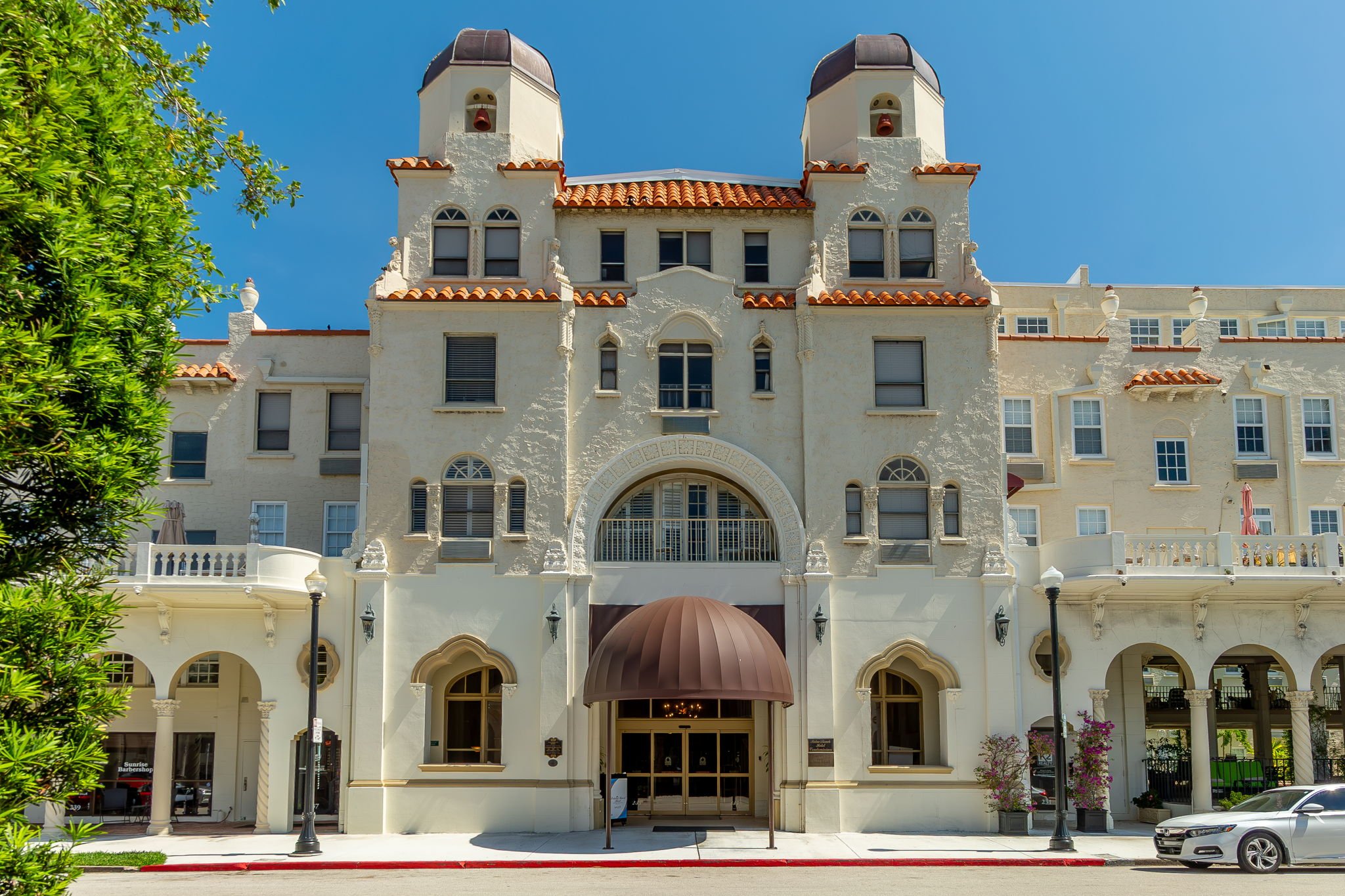 Front view of a large, ornate white building with a rounded brown awning over the main entrance, multiple arched and rectangular windows, and two prominent towers with turrets and small windows, under a clear blue sky, with some greenery and a parked