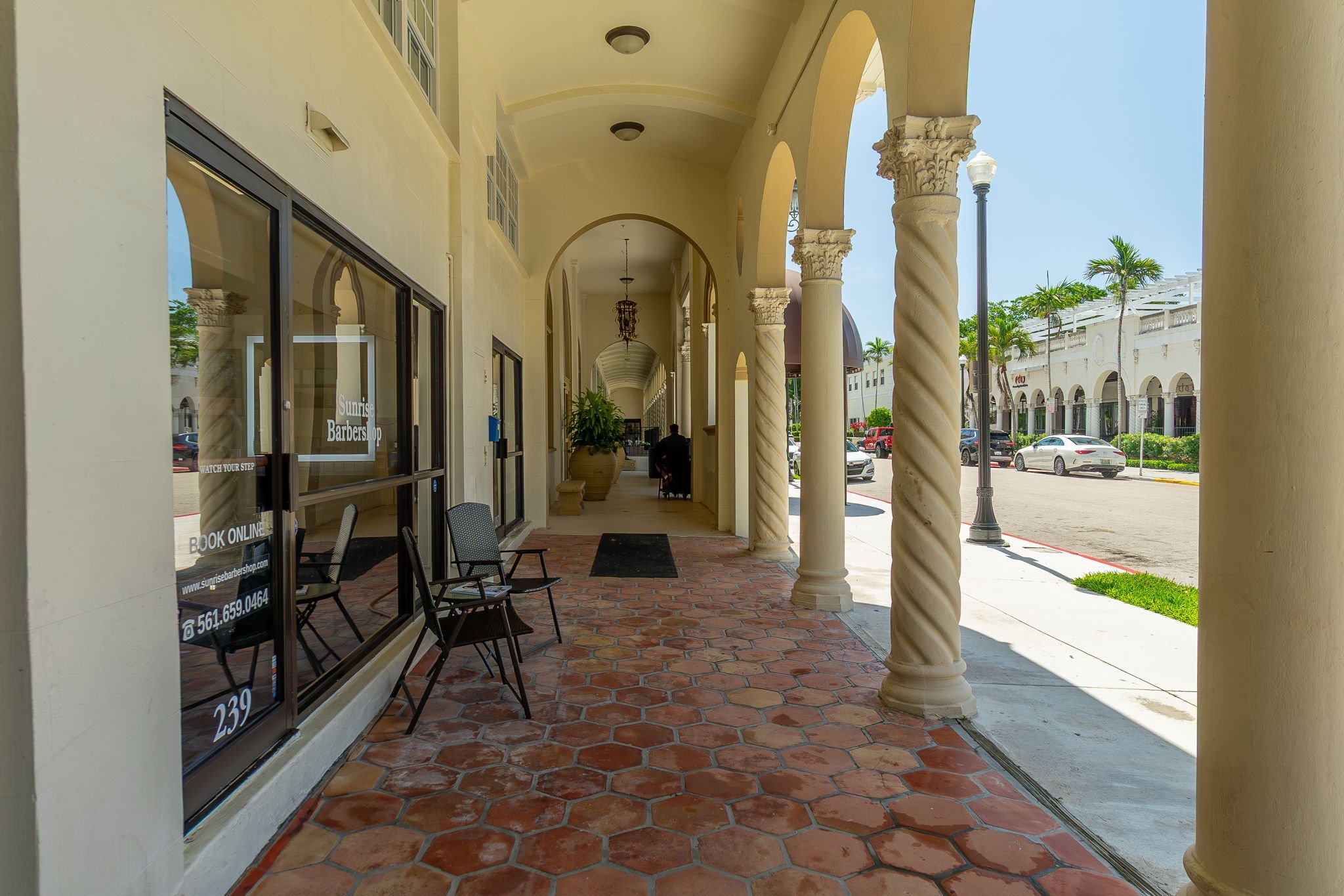 Outdoor shopping arcade with arched columns, brick tiled walkway, and storefronts on the left, parked cars on the street, and palm trees in the background.