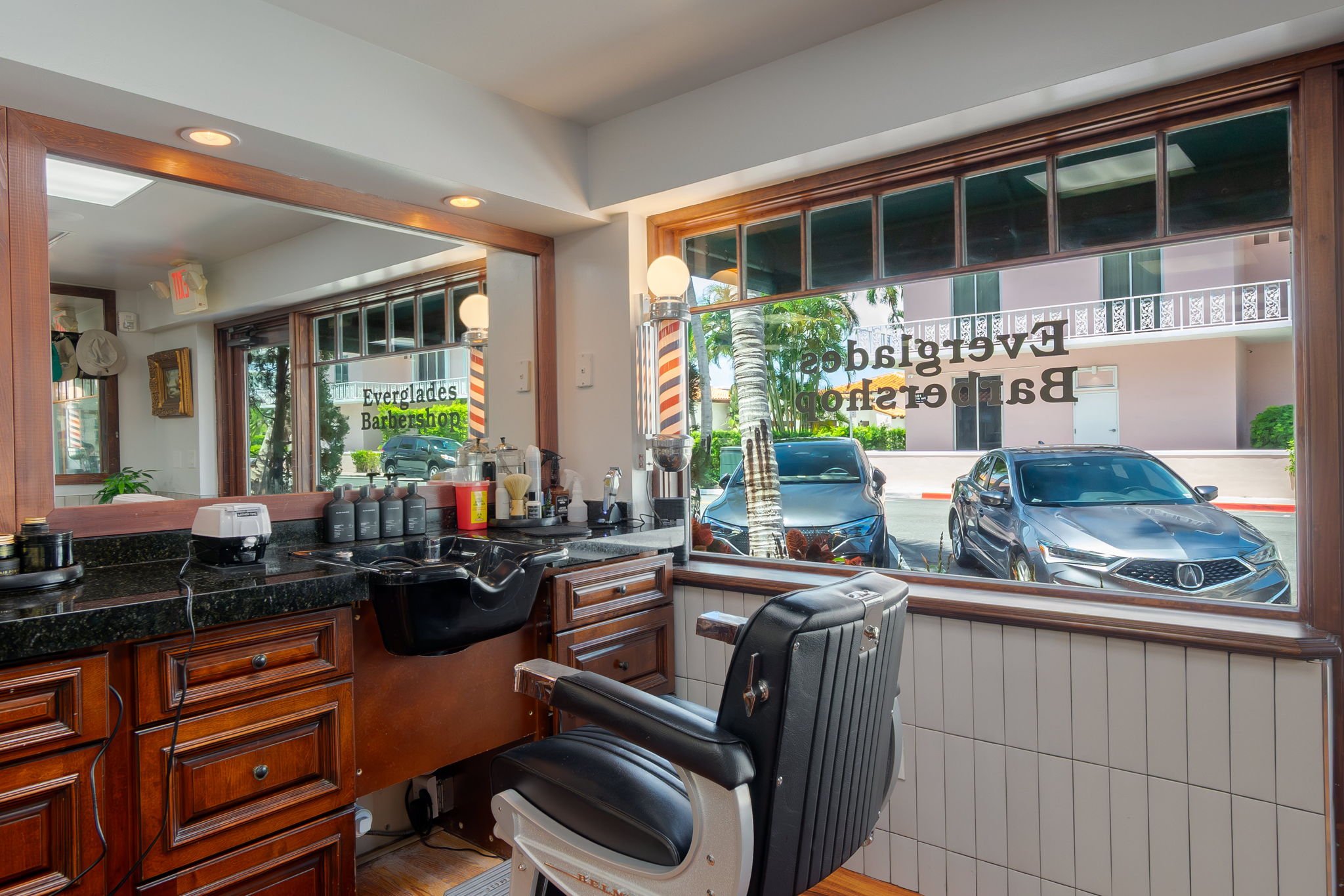 Inside a barbershop salon with a large front window showing parked cars outside, barber chairs, a mirror, and hair care products on the counter.