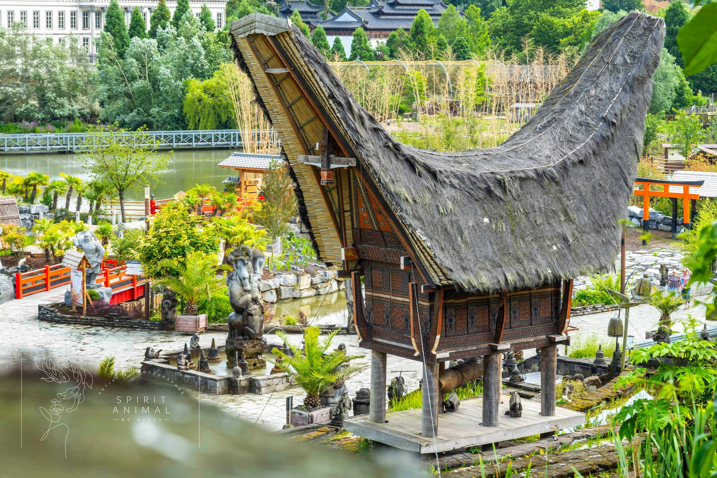 Traditionelles asiatisches Haus mit Strohdach, gebaut auf Holzpfählen, in einem gepflegten Garten mit Skulpturen, Pflanzen und Wasserlauf, umgeben von üppigem Grün, Fotografie von SPIRIT ANIMAL
