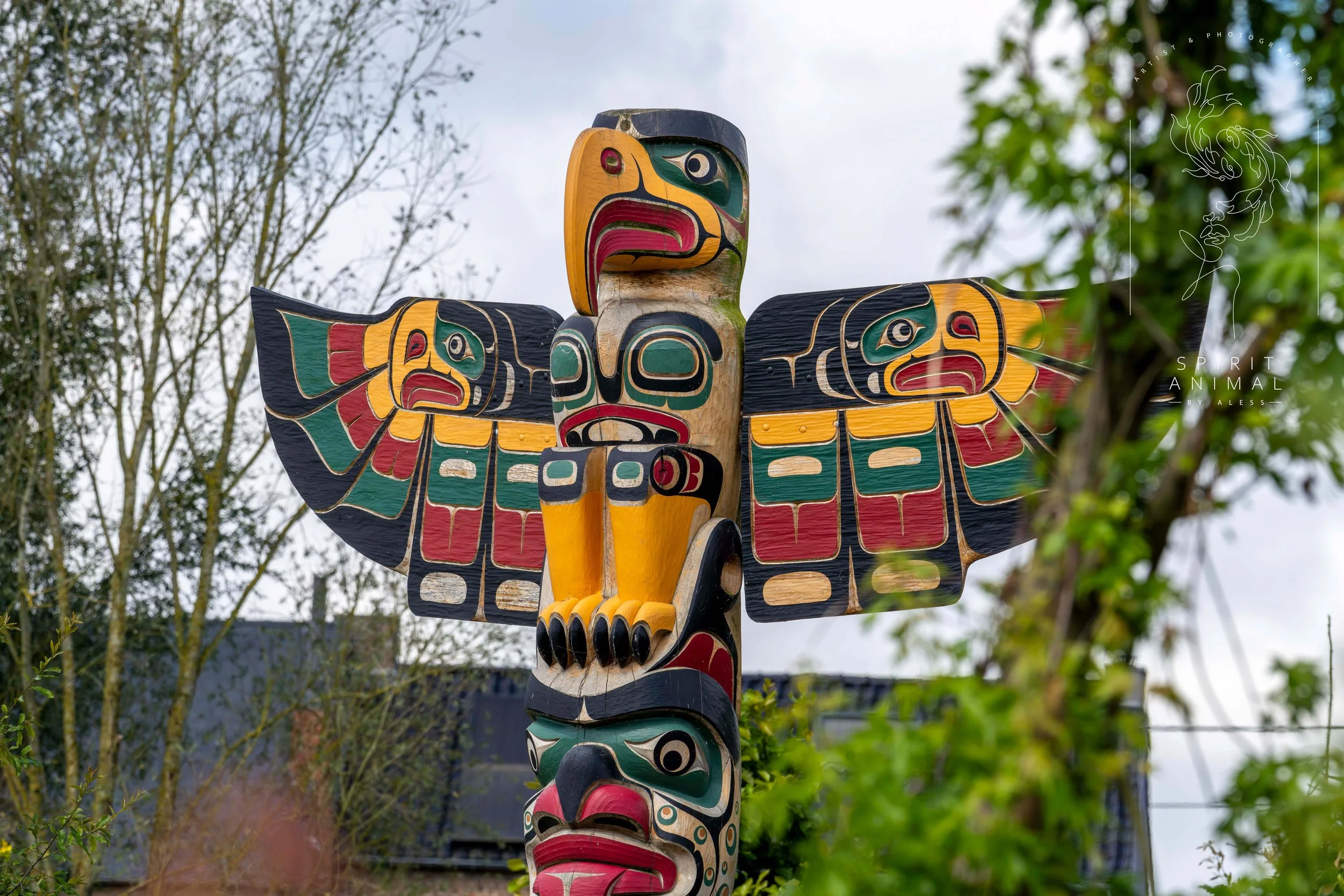 Ein bunter Totem-Polynesier-ähnlicher Holz-Skulptur mit Tier- und Menschengesichtern, vor Bäumen und einem Himmel mit Wolken, Fotografie von SPIRIT ANIMAL