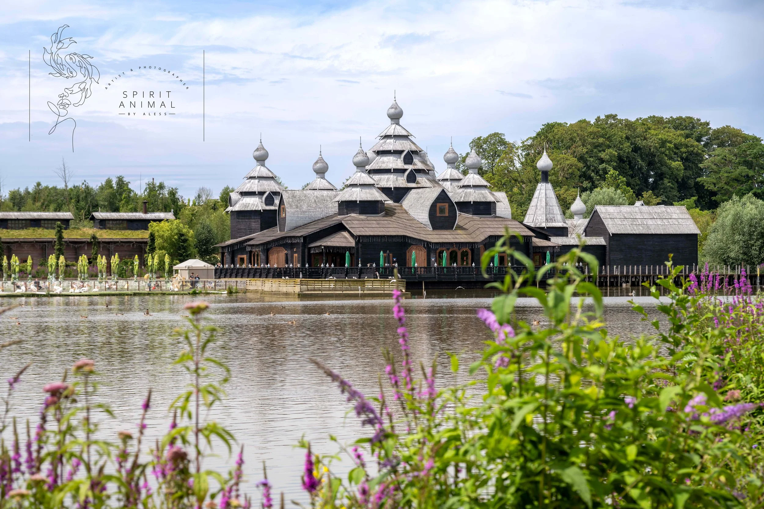 Ein dunkles, geschnitztes Holzgebäude mit mehreren silbernen, spitzen Türmen, gelegen an einem See mit Blumen im Vordergrund und Bäumen im Hintergrund, Izzba Pairi Daiza, Fotografie von SPIRIT ANIMAL