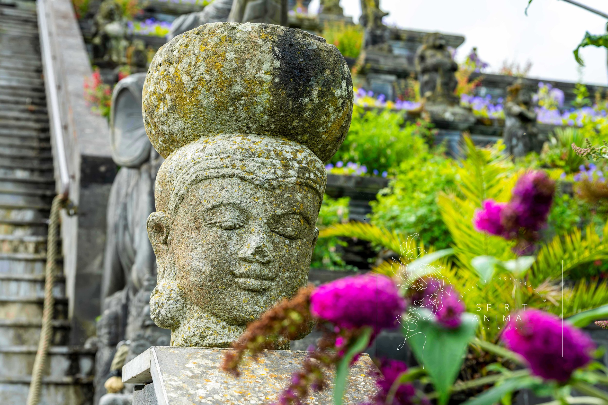 Steinfigur eines Gesichts mit geschlossenen Augen, umgeben von bunten Blumen im Garten, Fotografie von SPIRIT ANIMAL